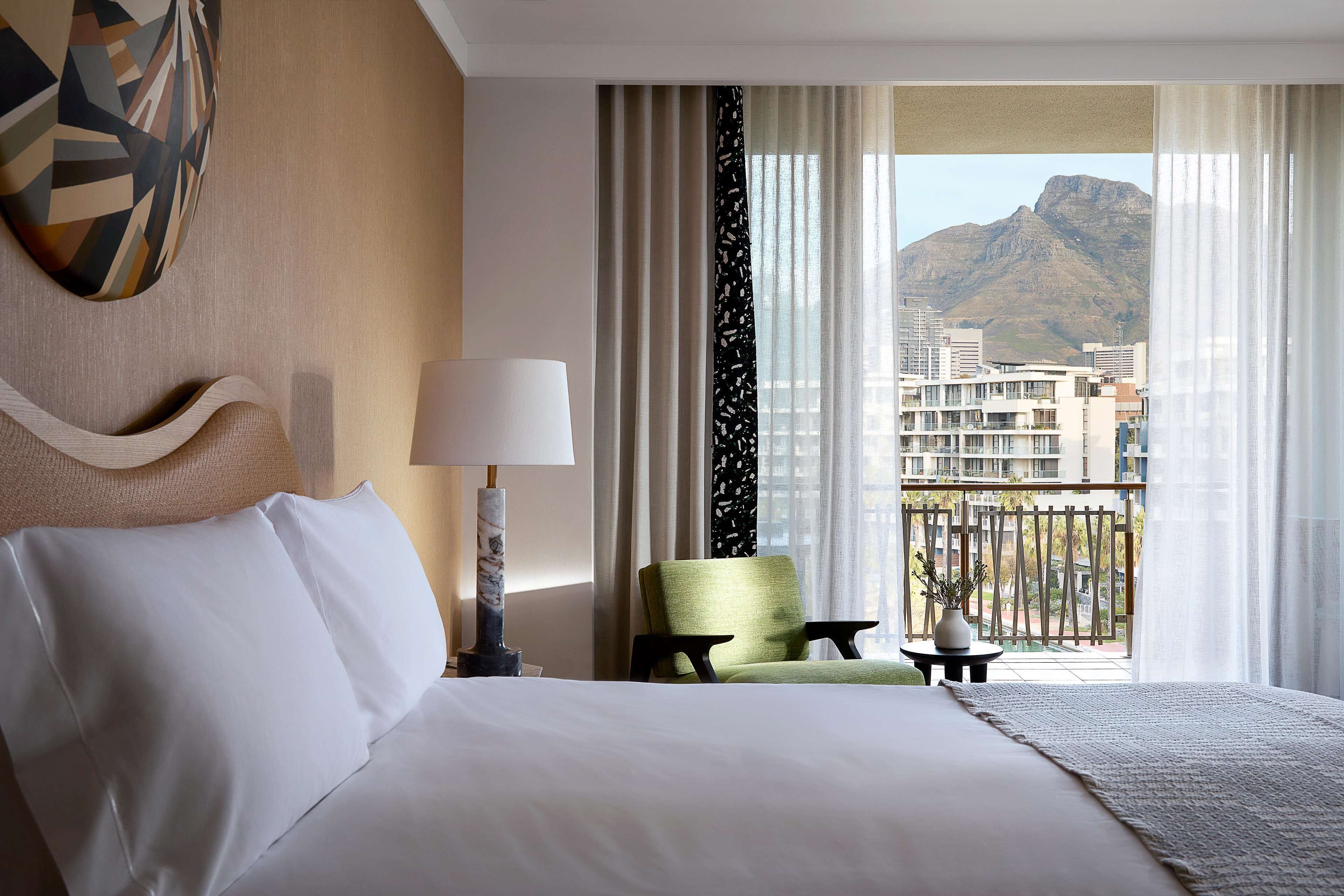 A hotel room with a large bed, a green armchair, a small table with a vase, and a view of buildings and mountains through sheer curtains.
