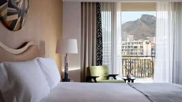 A hotel room with a large bed, a green armchair, a small table with a vase, and a view of buildings and mountains through sheer curtains.