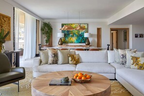 A modern living room with a sectional sofa, a round coffee table with a book and a bowl of fruit, and a large abstract painting above the sofa.
