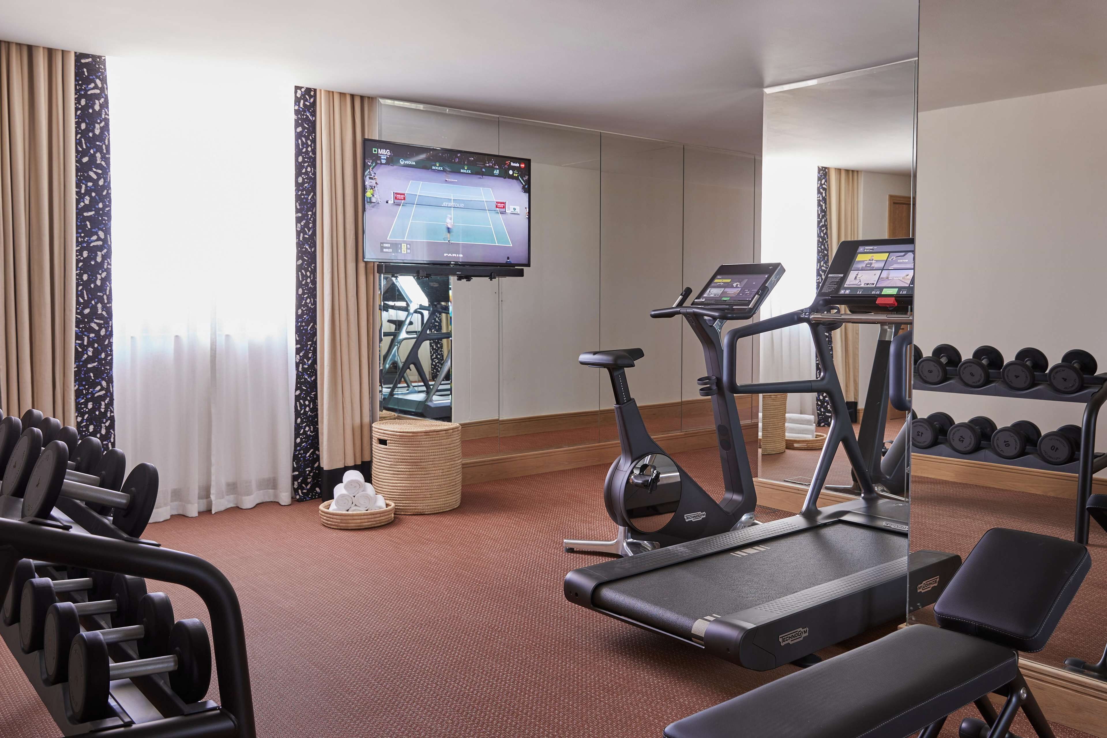 A hotel gym with treadmills, dumbbells, and a TV displaying a tennis match.