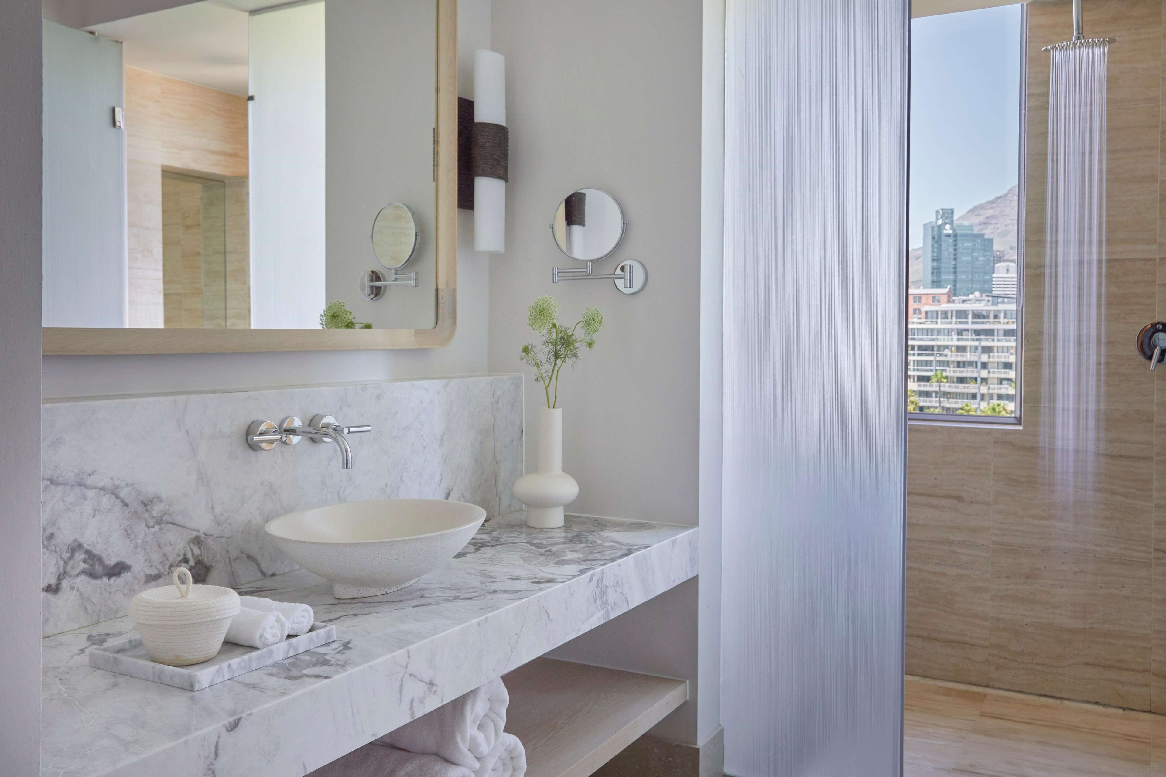 A modern bathroom with a marble sink, a large mirror, a vase with greenery, and a view of a cityscape through a window.