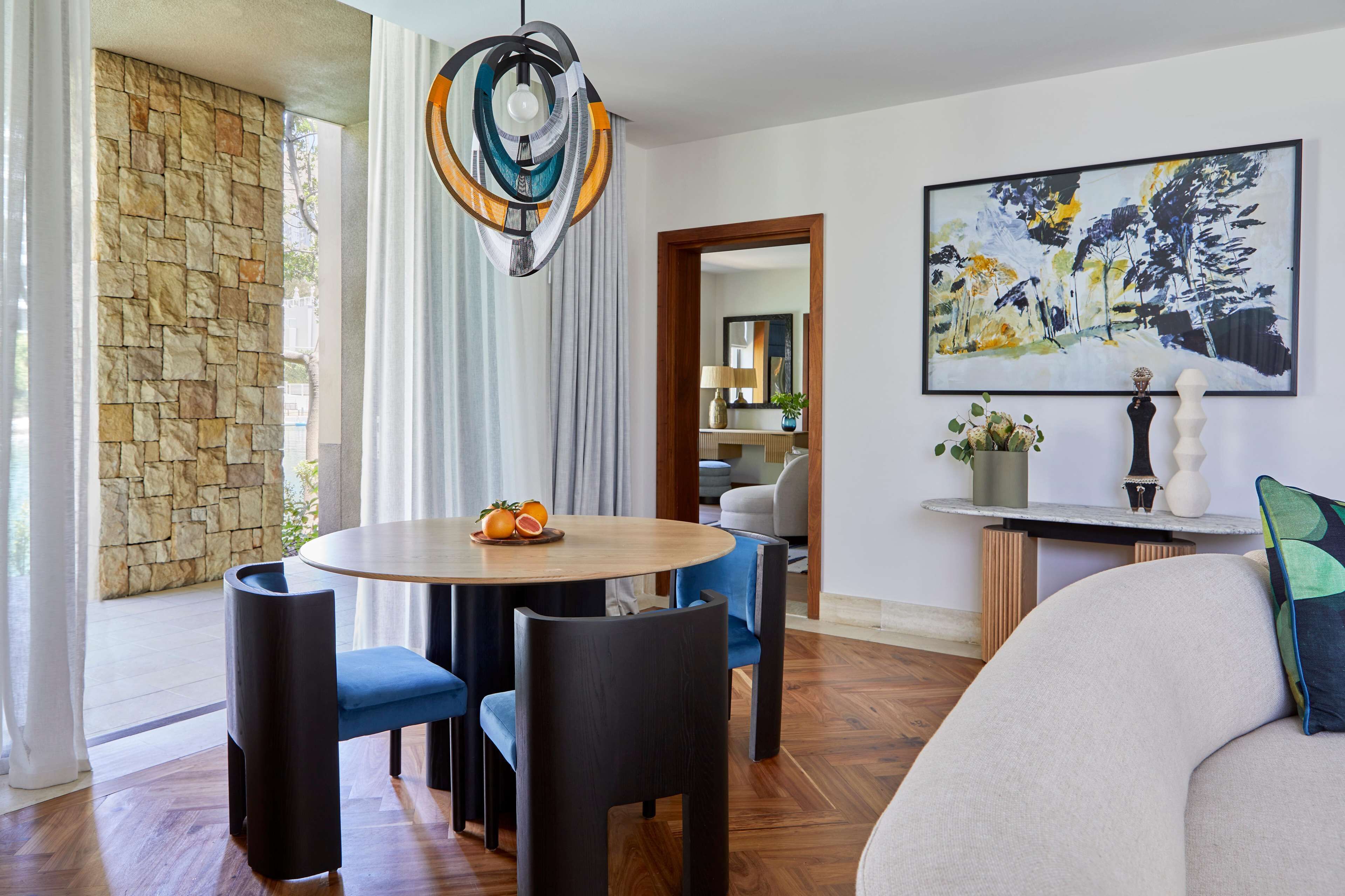 A modern living room with a stone wall, a round wooden table with fruit, blue chairs, a sofa, and a large abstract painting.