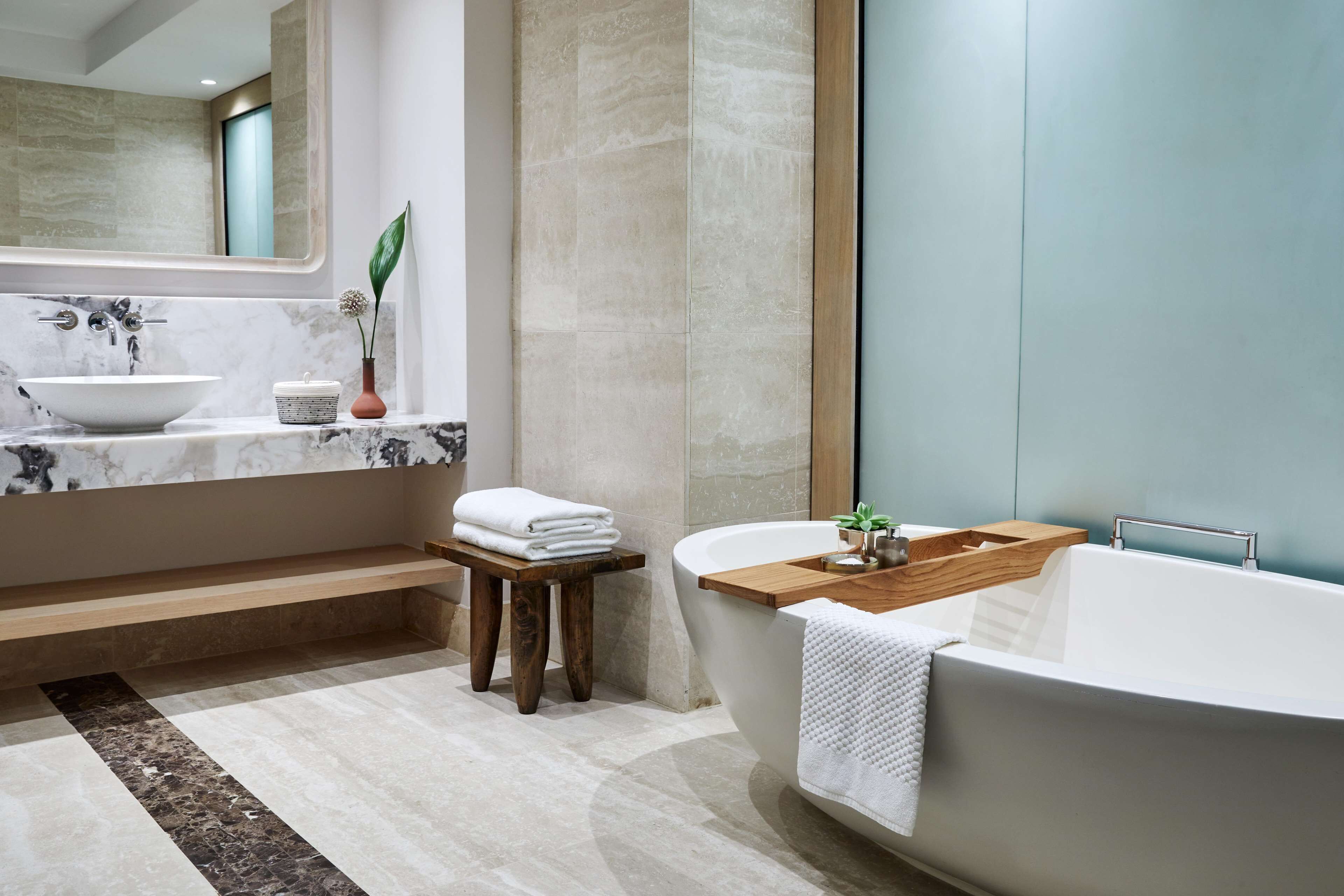 Un baño moderno con una bañera independiente, un tocador de mármol y un taburete de madera.