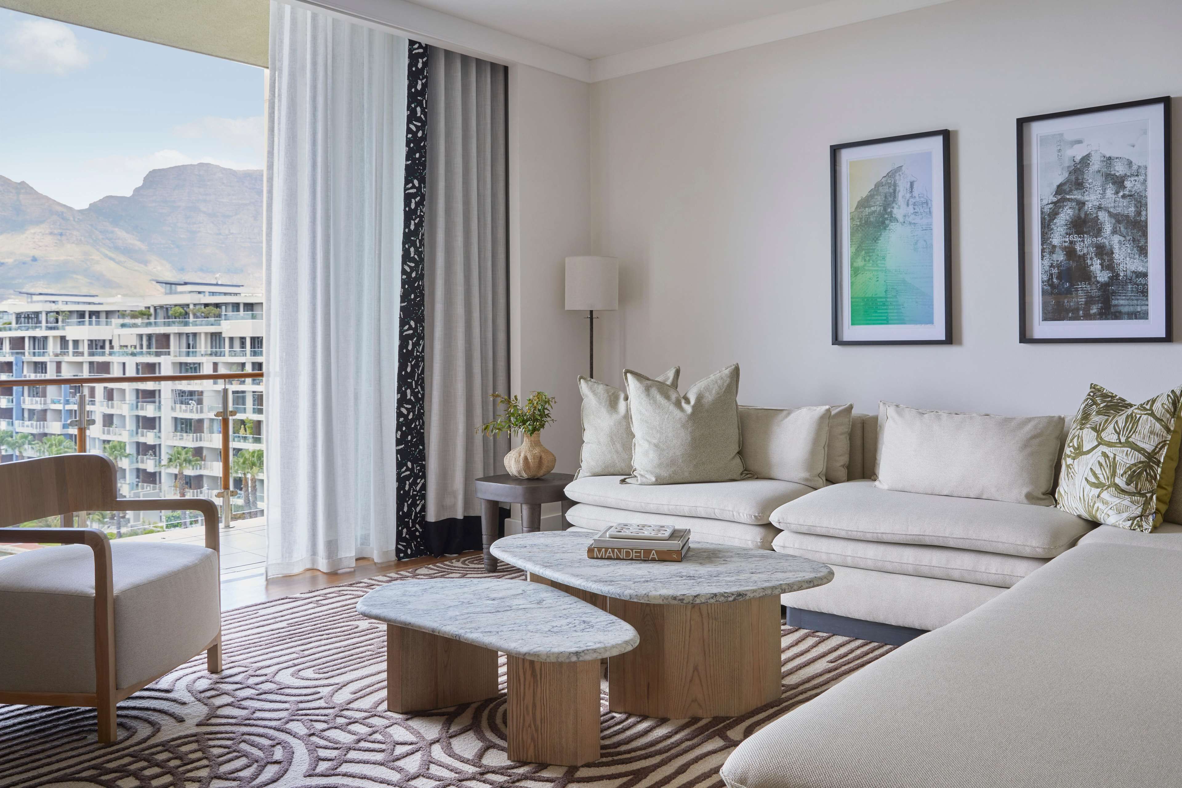 A modern hotel room with a large window offering a mountain view, a sofa set, a coffee table, and a lamp.