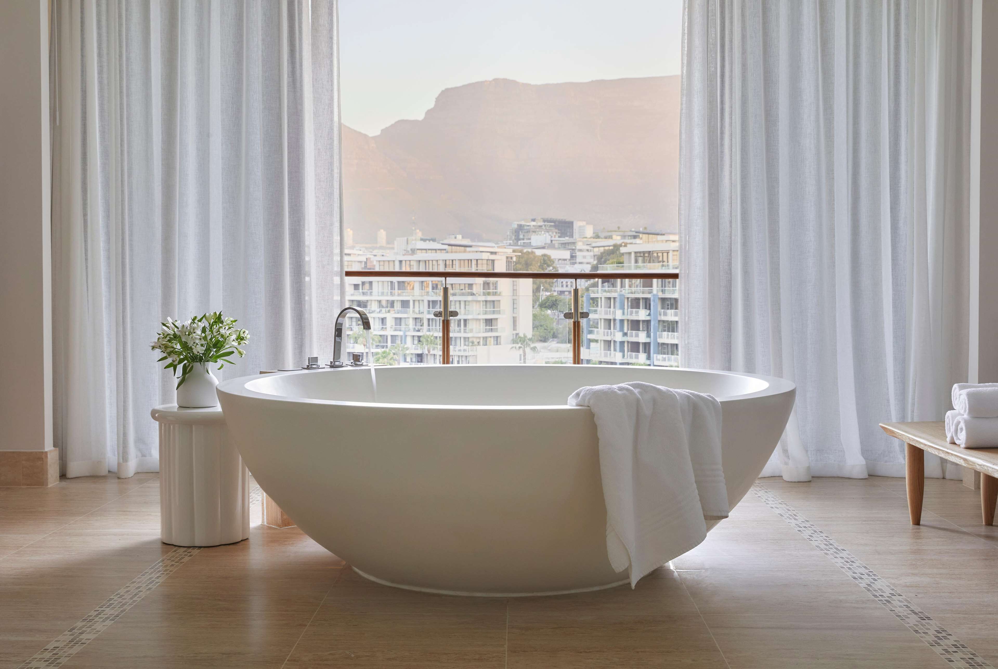 Un baño moderno con una bañera independiente, una mesita auxiliar con una planta y vistas a un paisaje urbano a través de grandes ventanales.