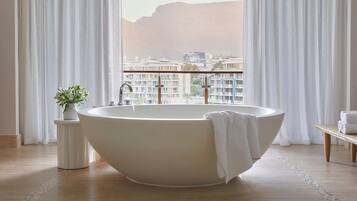 Un baño moderno con una bañera independiente, una mesita auxiliar con una planta y vistas a un paisaje urbano a través de grandes ventanales.
