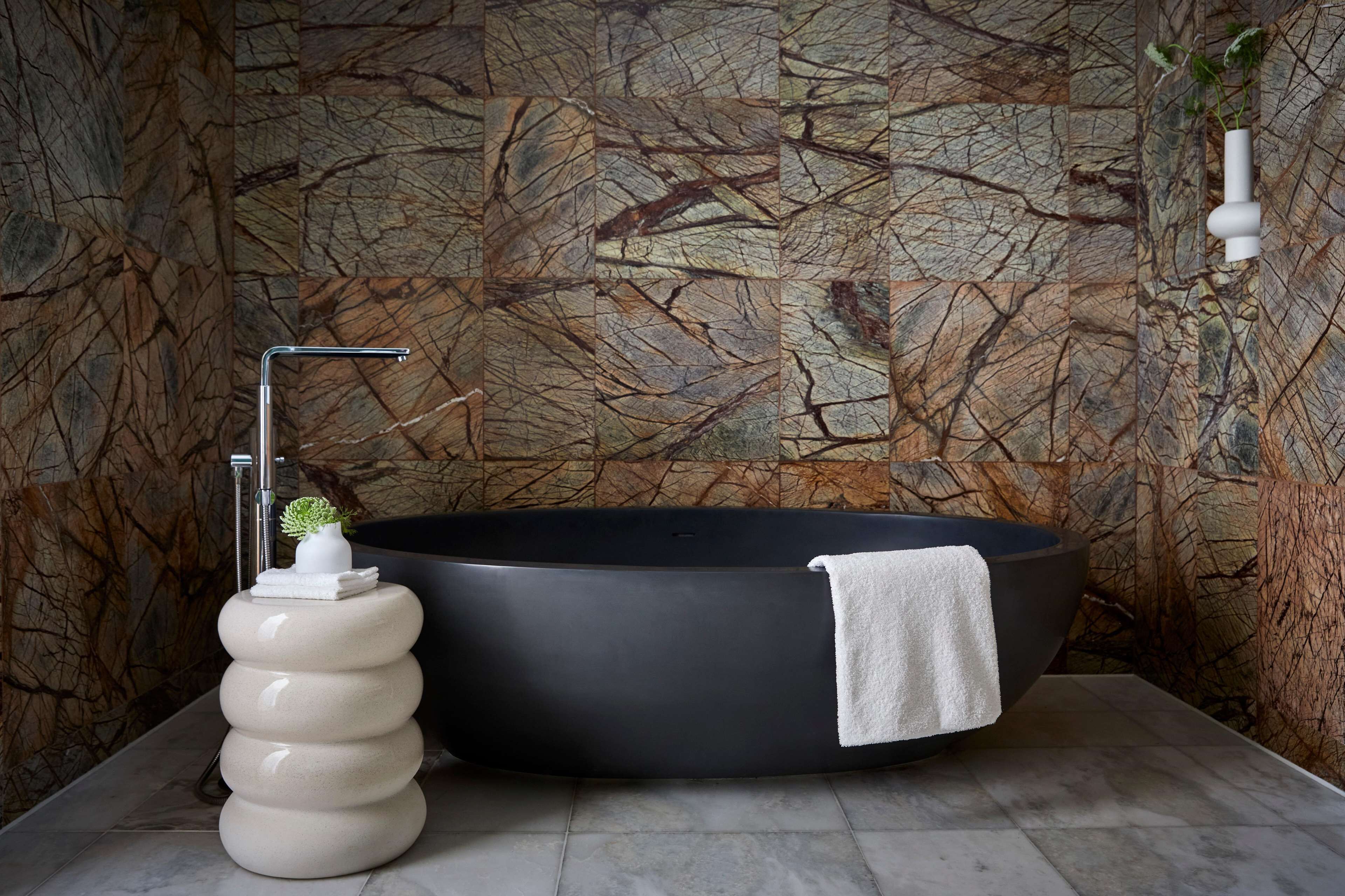 A modern bathroom with a large black bathtub, a white marble pedestal, and a textured wall.