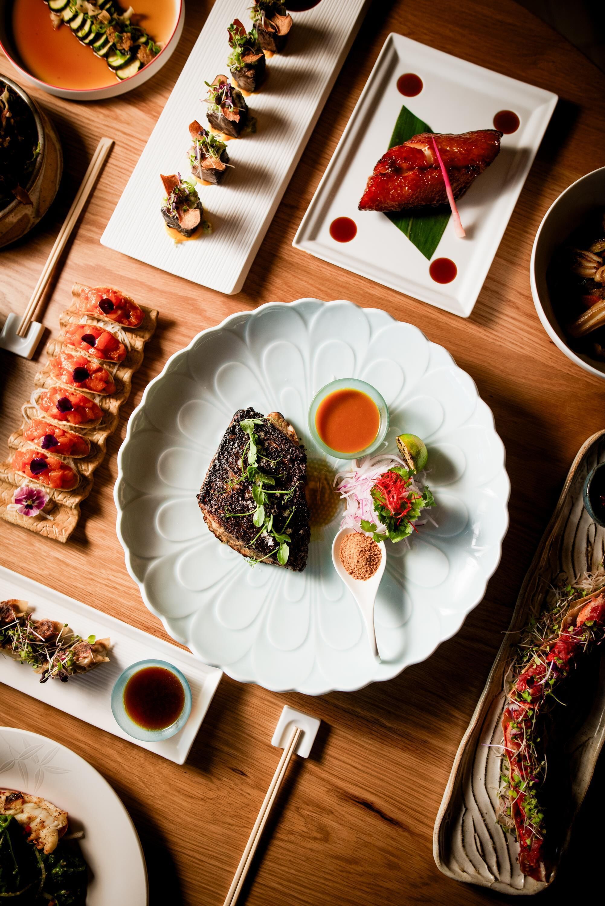 Une variété de plats sont disposés sur une table en bois, notamment des sushis, des sashimis et d’autres amuse-gueule, accompagnés de baguettes et de petits bols de sauce.