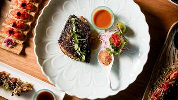 Une variété de plats sont disposés sur une table en bois, notamment des sushis, des sashimis et d’autres amuse-gueule, accompagnés de baguettes et de petits bols de sauce.