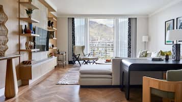 A modern living room with a wooden floor, a sofa, a dining table, and a TV. There is a view of mountains through large windows.