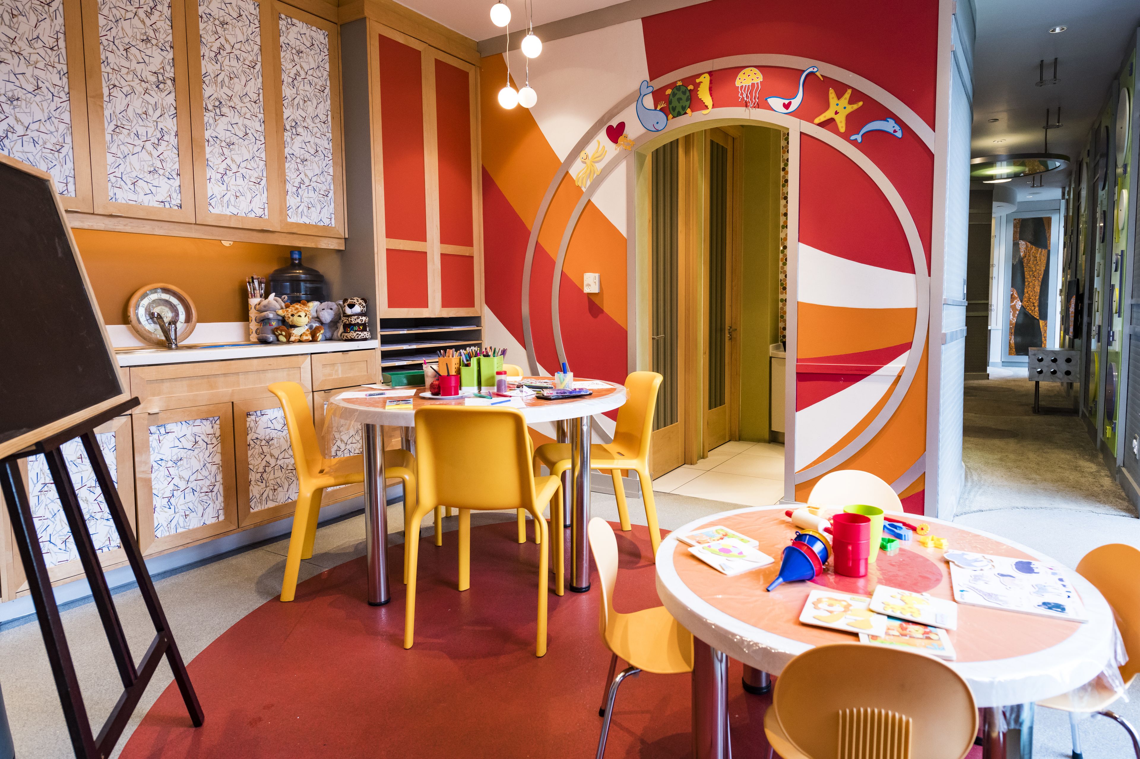 Une salle de jeux pour enfants aux couleurs vives, avec des chaises et des tables jaunes, une grande décoration murale circulaire et un meuble en bois doté de portes en verre à motifs.