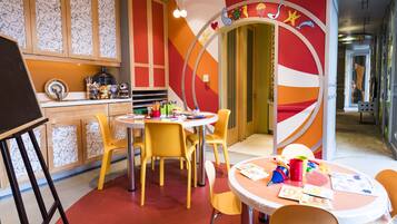 A brightly colored children's playroom with yellow chairs and tables, a large circular wall decoration, and a wooden cabinet with patterned glass doors.