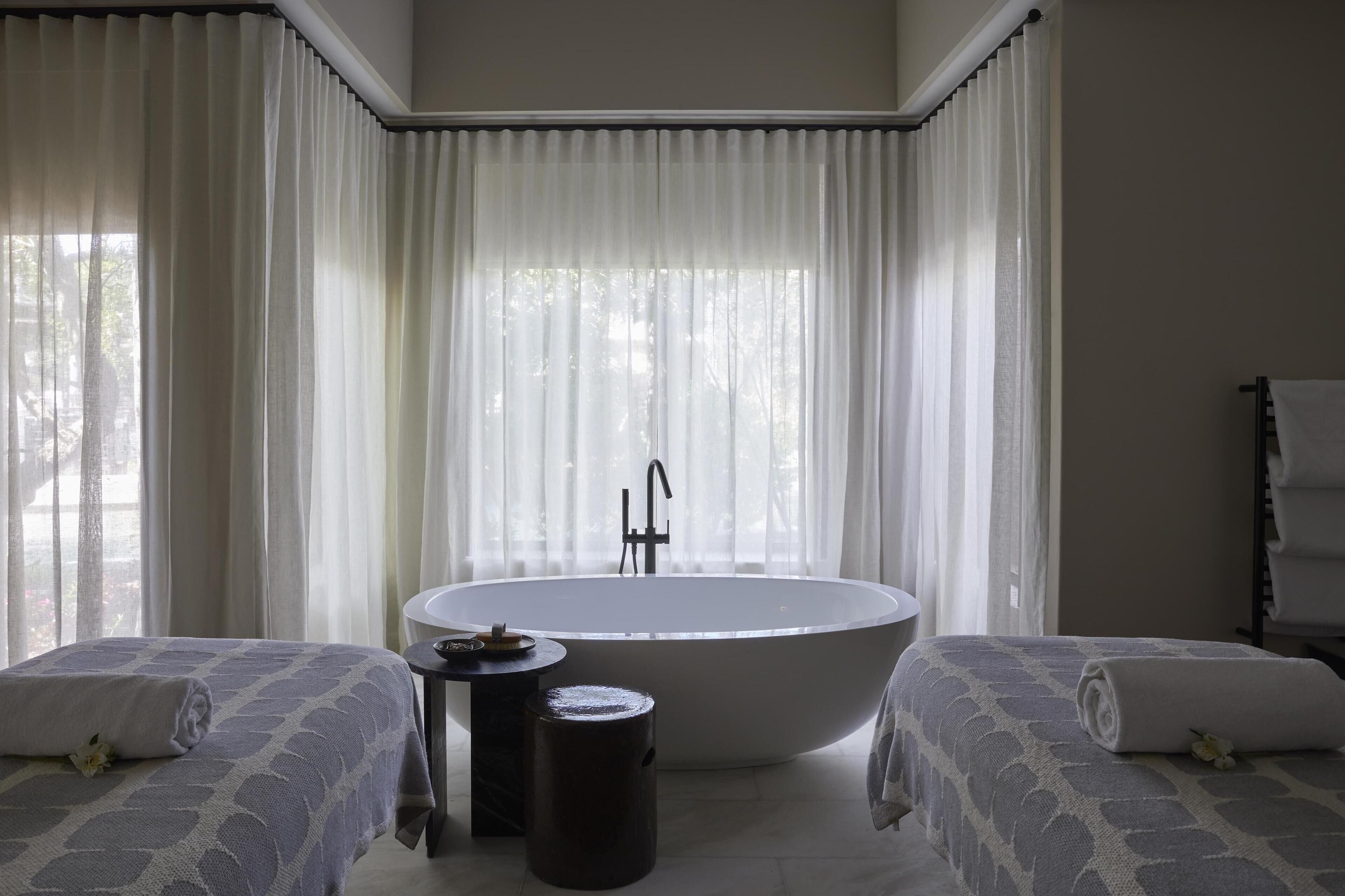 Un baño moderno con una bañera independiente, dos camas con toallas, una mesita redonda pequeña y cortinas de gasa.