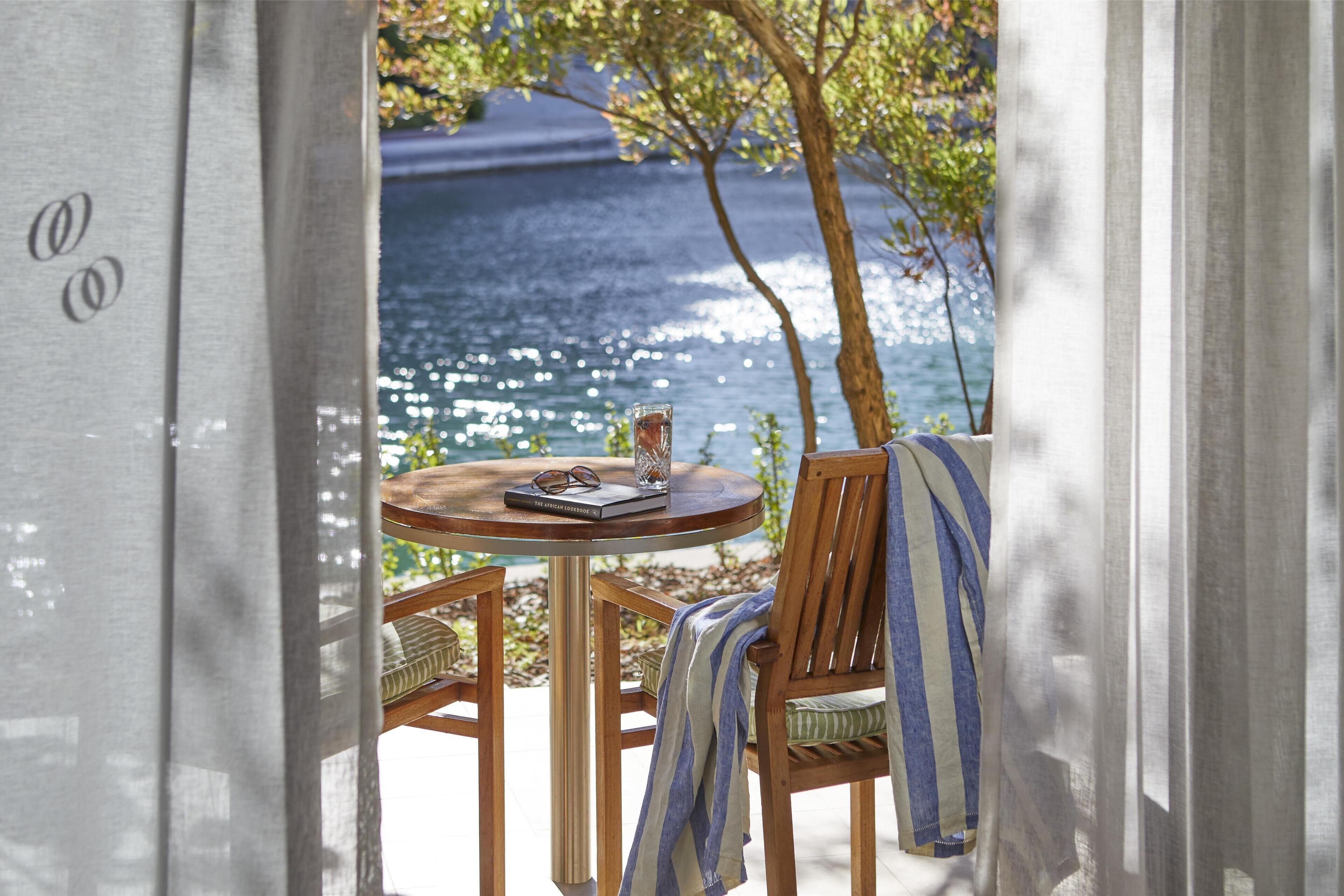 A wooden table with a book and glasses, a chair with a striped towel, and a view of the ocean through a window.