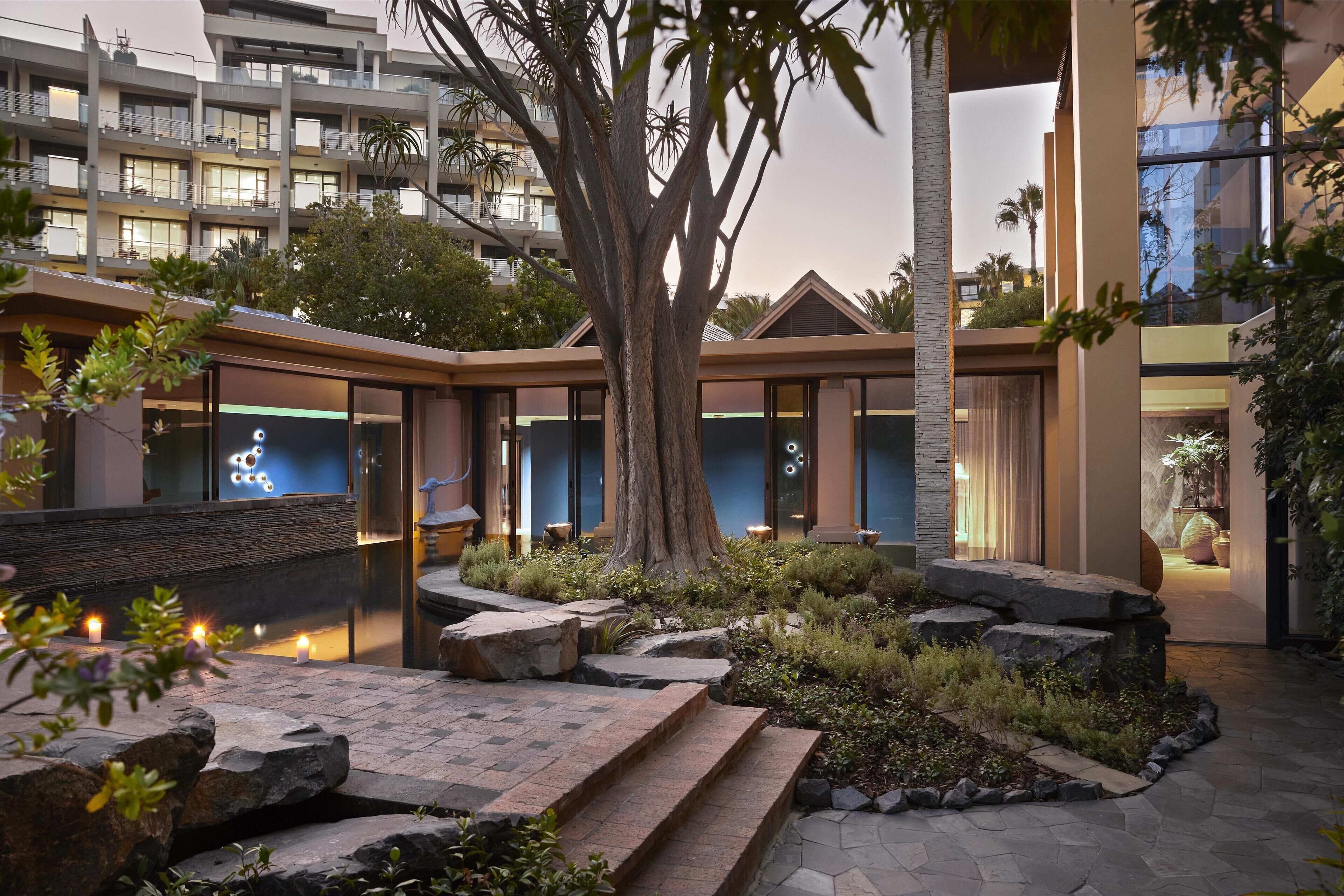Une cour d’hôtel moderne avec un grand arbre, des allées pavées et un espace piscine.