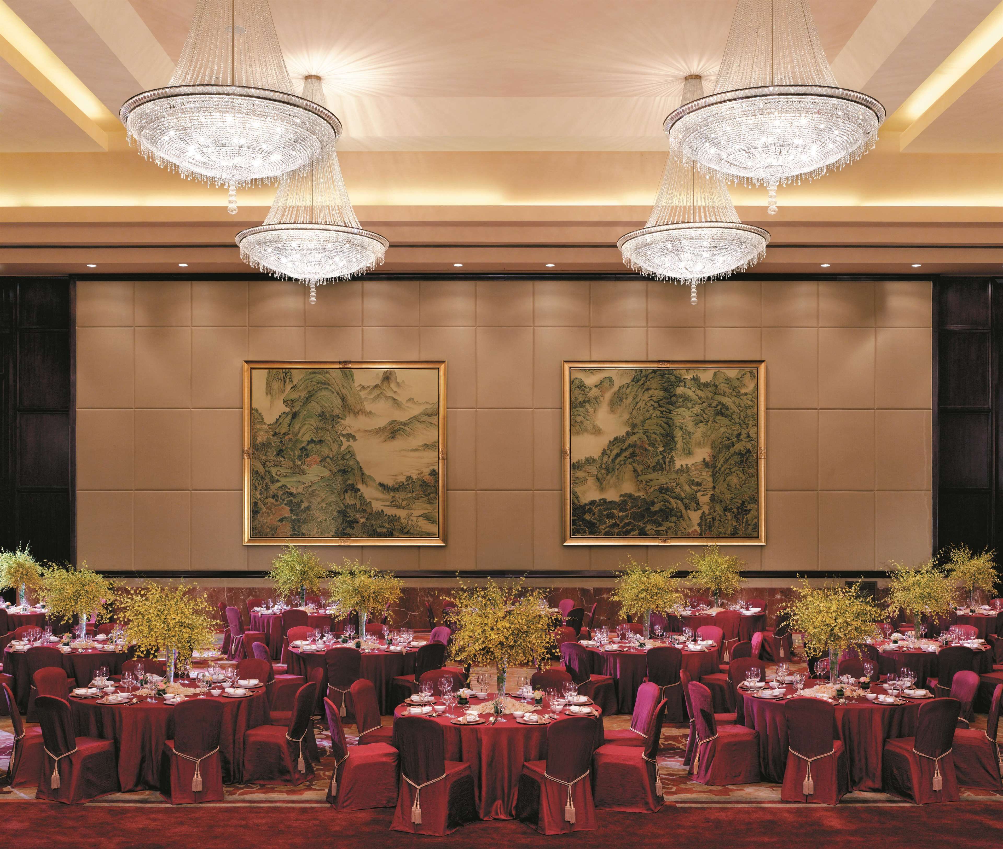 A banquet hall with round tables set for dining, large chandeliers, and traditional paintings on the walls.