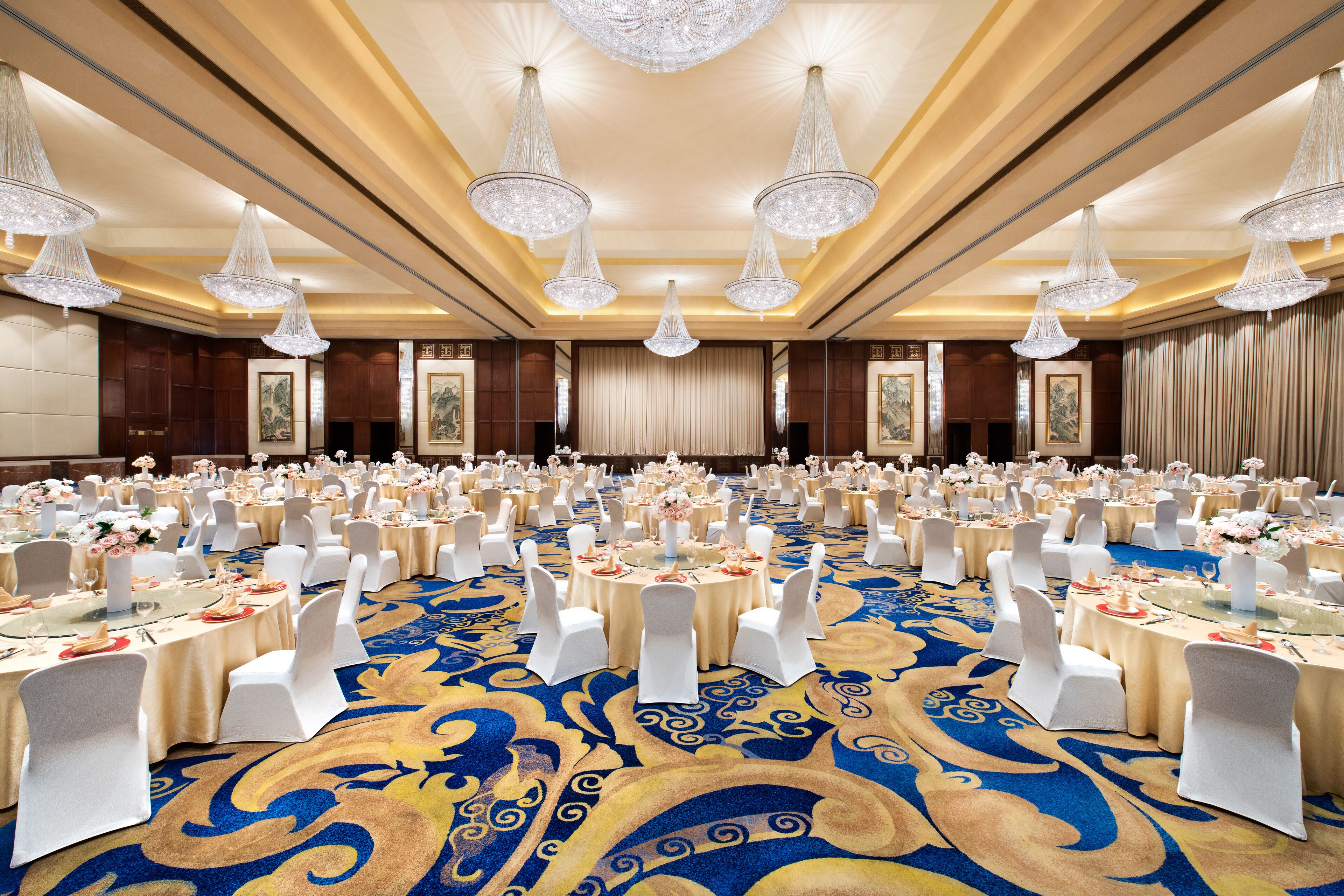A banquet hall with round tables, white chairs, and chandeliers.