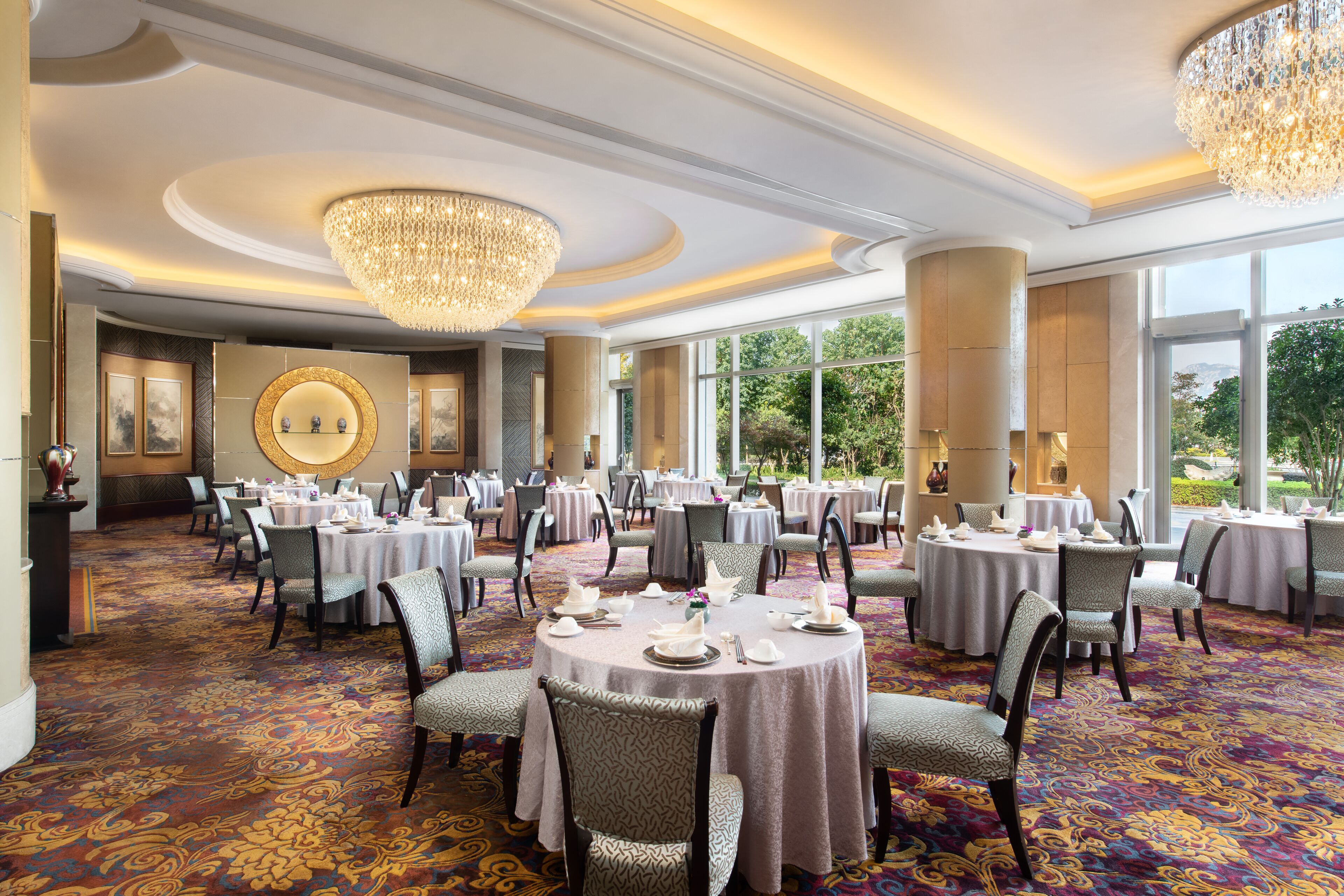 Un amplio salón de banquetes con mesas redondas, elegantes candelabros y suelo alfombrado.