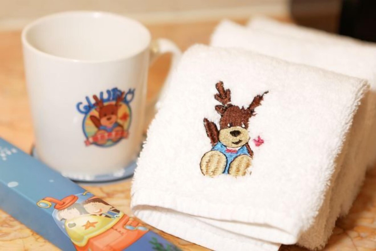 A white mug with a colorful cartoon character, a white towel with a teddy bear embroidered on it, and a blue card with a cartoon character.