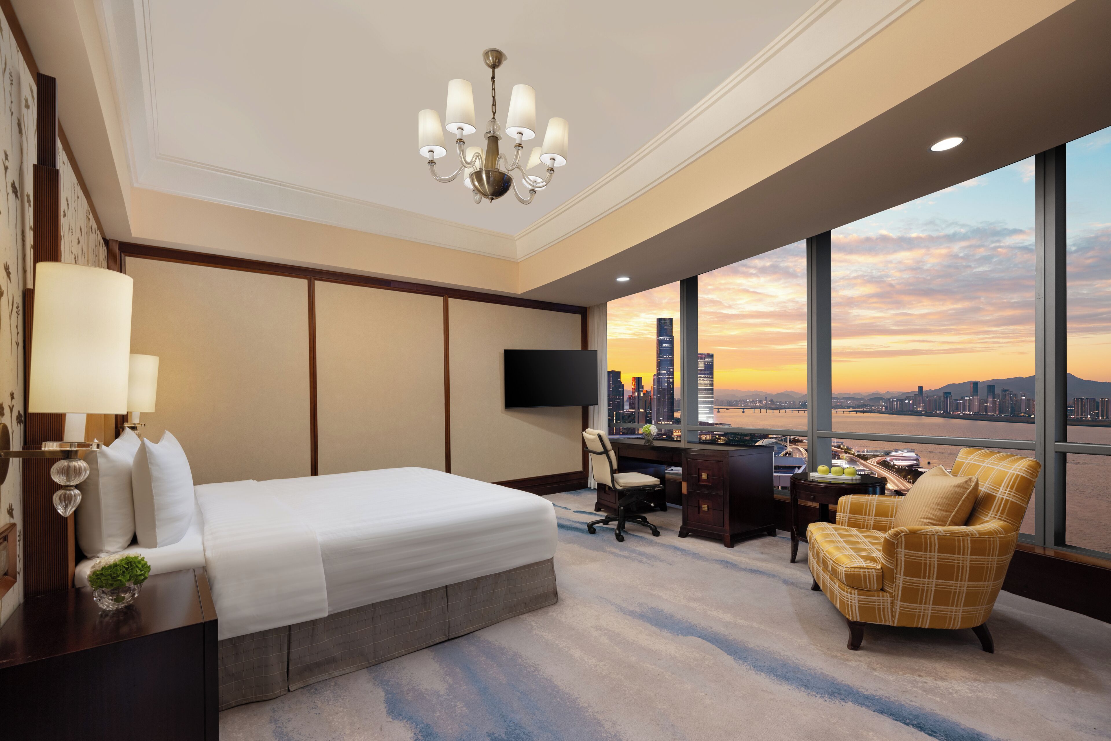 Habitación de hotel con una cama grande, un escritorio, una silla, un televisor y vistas del horizonte urbano al atardecer.
