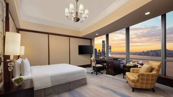 Habitación de hotel con una cama grande, un escritorio, una silla, un televisor y vistas del horizonte urbano al atardecer.