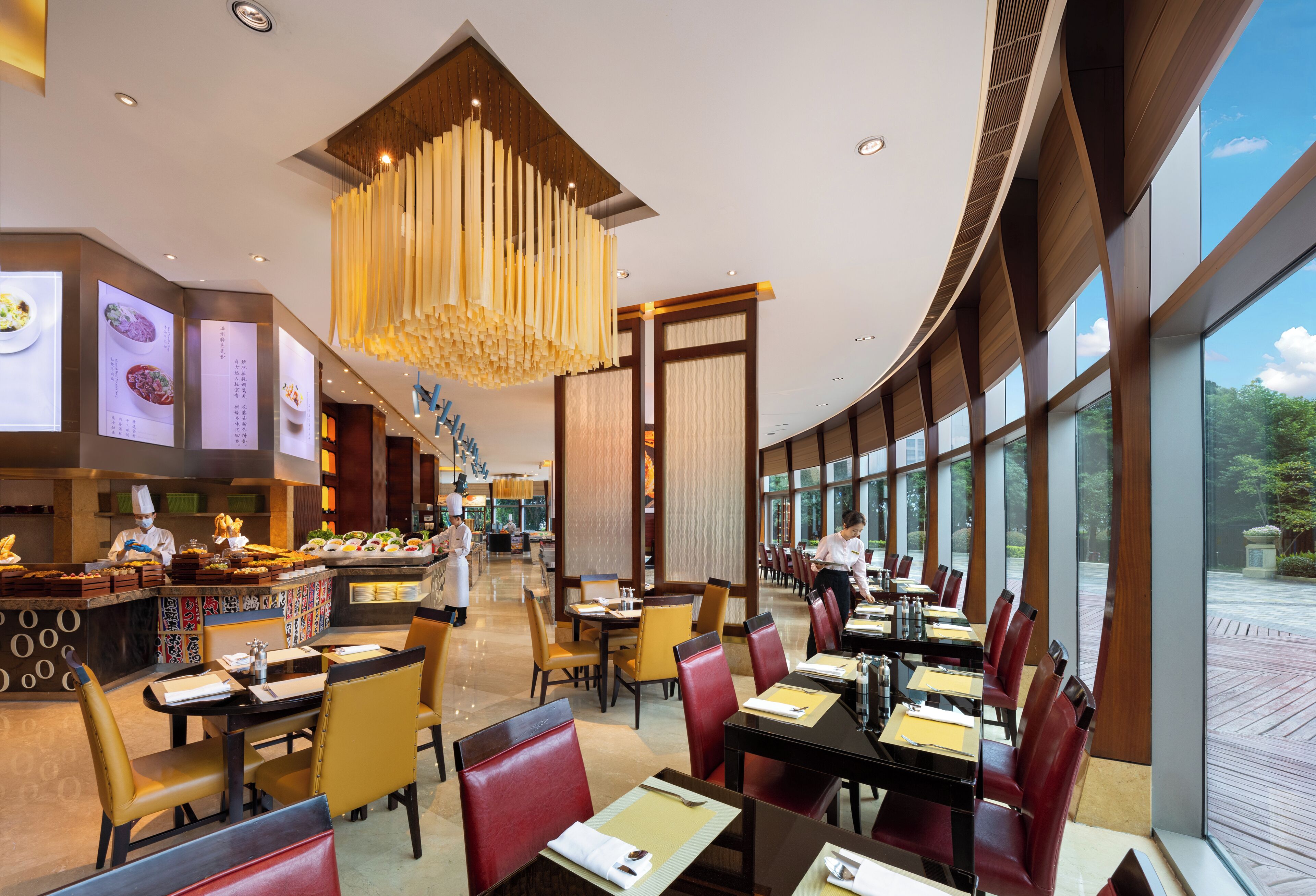 A modern restaurant with a central bar, high ceilings, and large windows offering views of the outdoors.