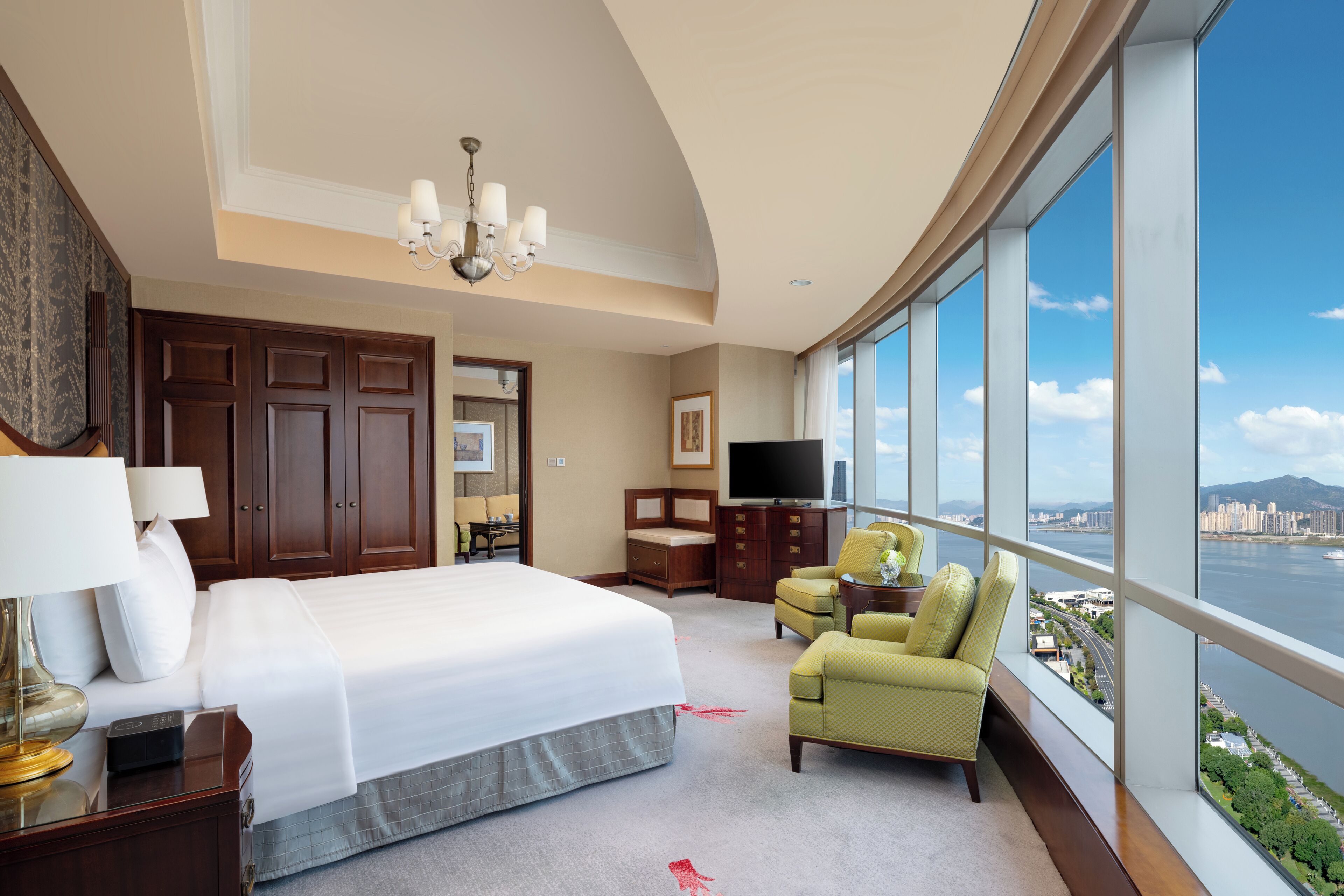 A hotel room with a large bed, a television, a desk with a lamp, a chair, and a view of a city and water.