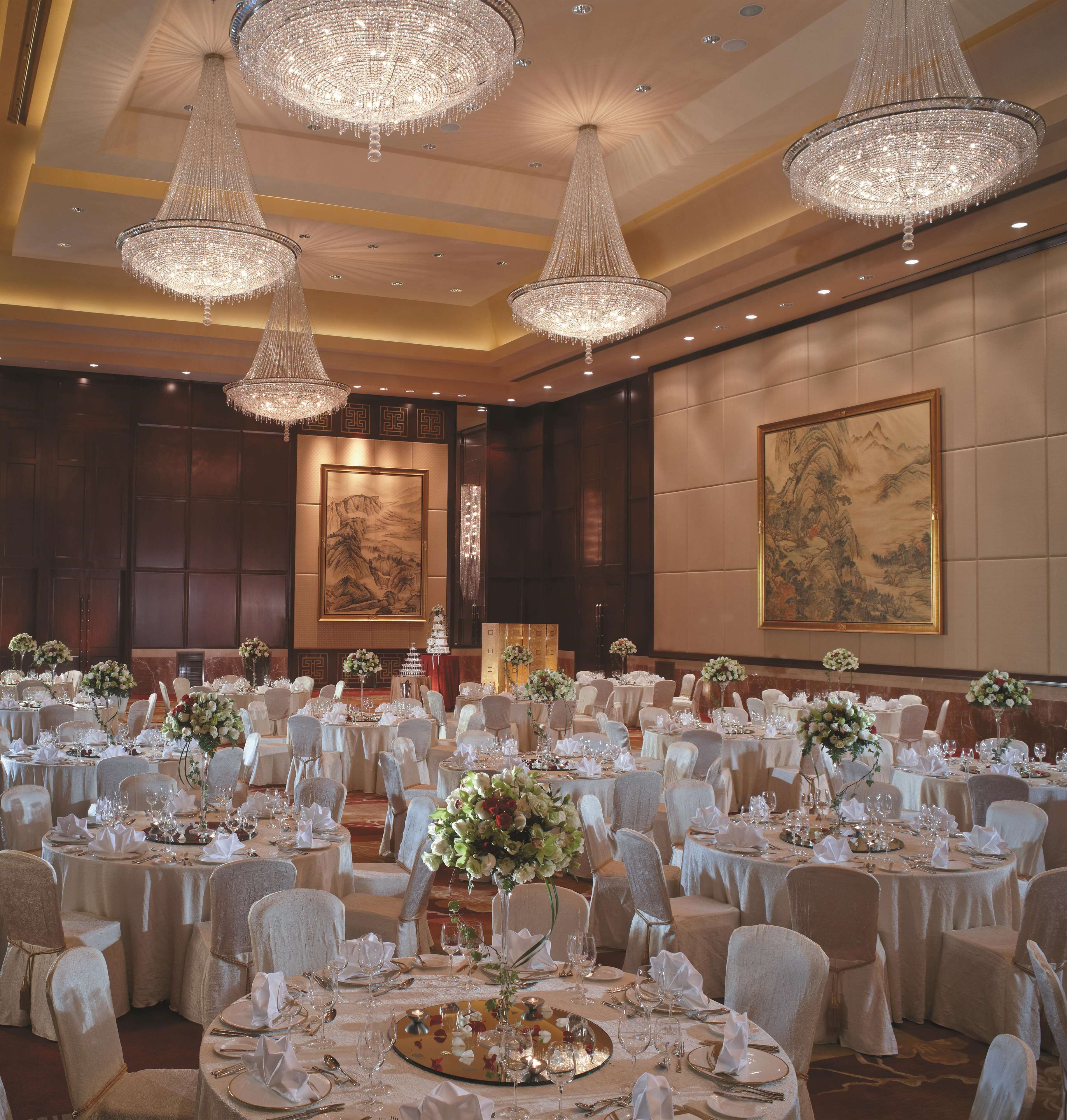 Een balzaal met ronde tafels gedekt voor een formeel evenement, grote kroonluchters en traditionele kunstwerken aan de muren.