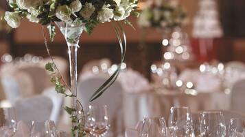 Mesa posta para um evento formal, com um arranjo floral alto, copos de vidro transparente e toalhas de mesa brancas.