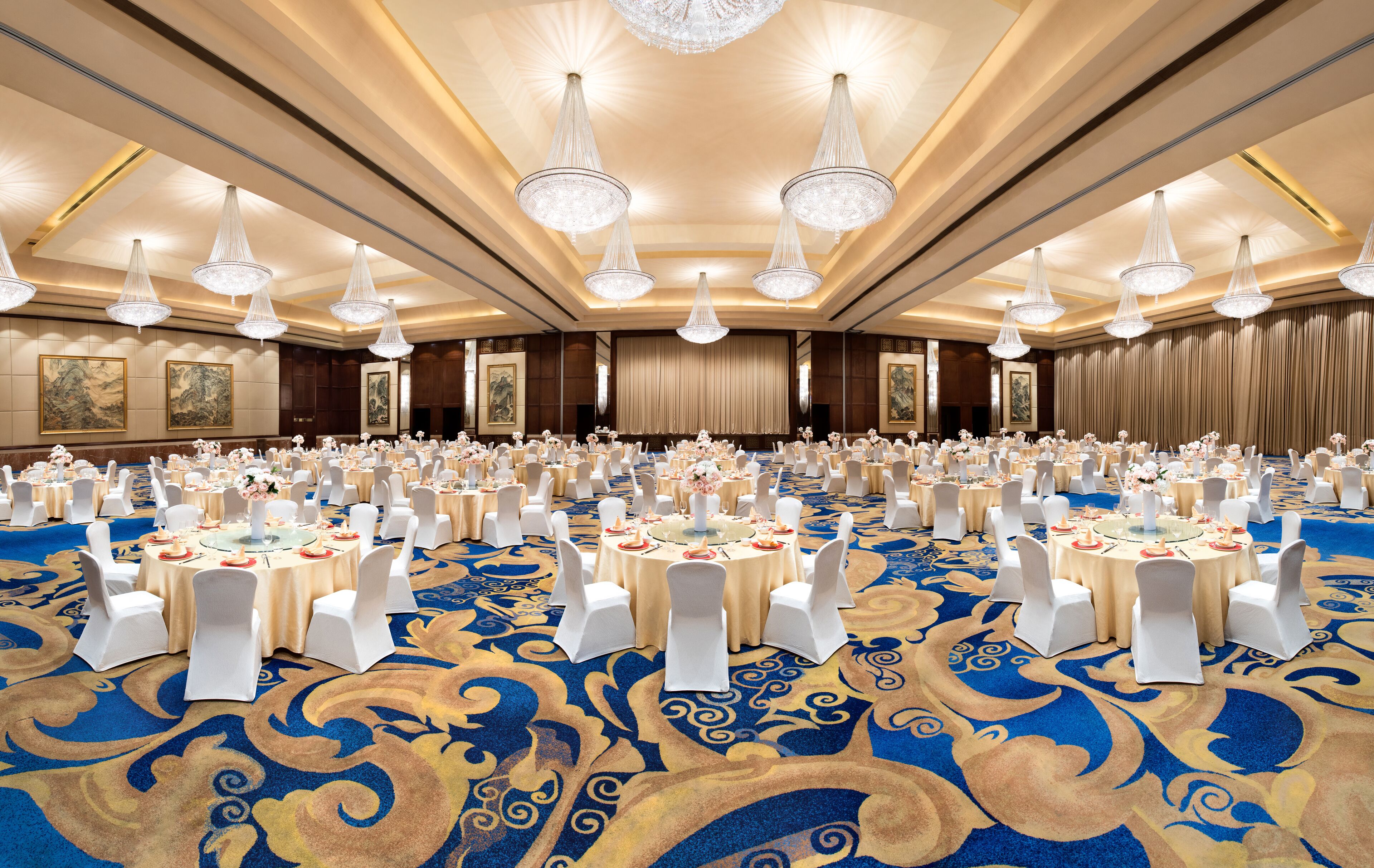 A large banquet hall with round tables, white chairs, and chandeliers.