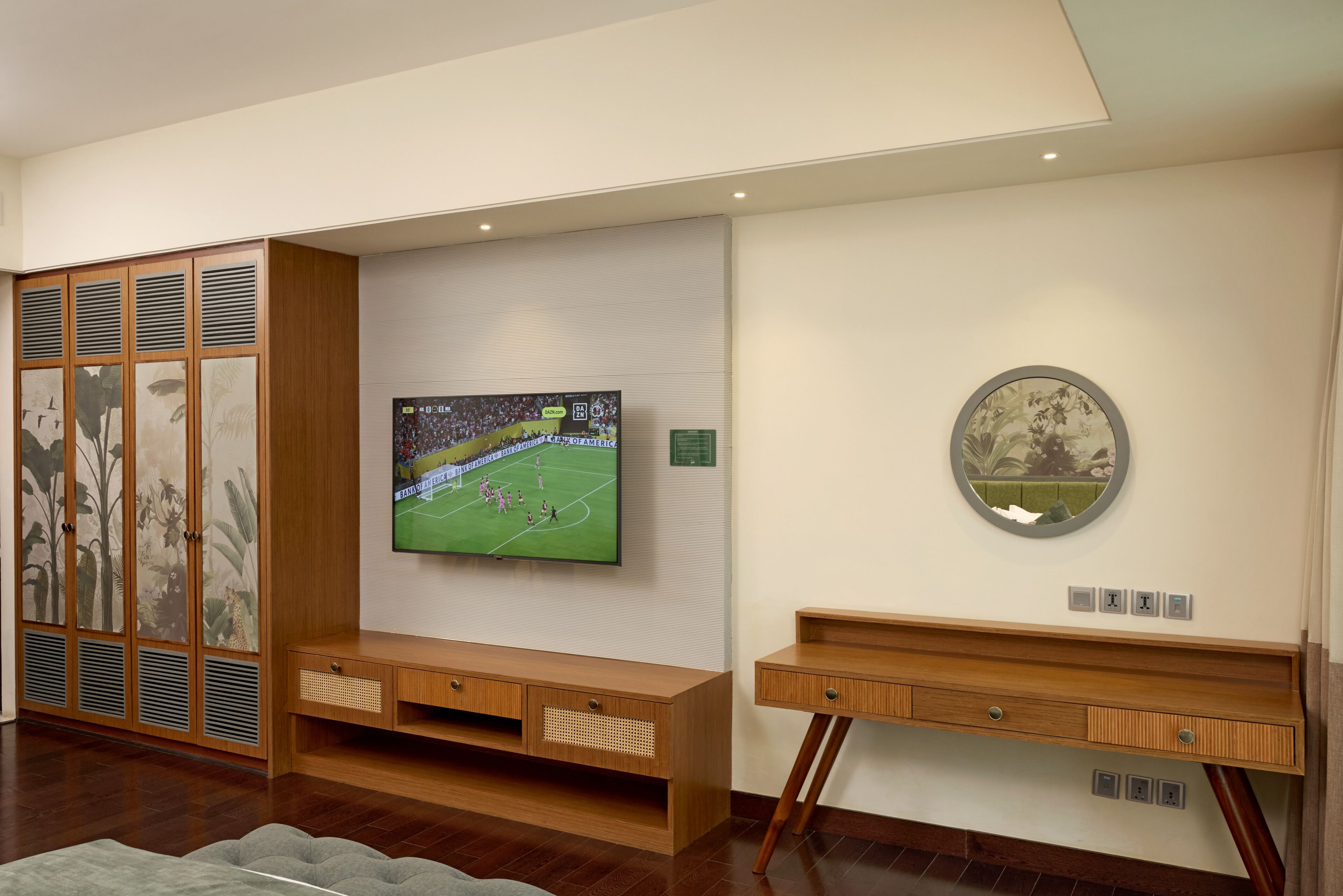 A modern living room with a flat-screen TV mounted on the wall, wooden furniture, and a decorative mirror.