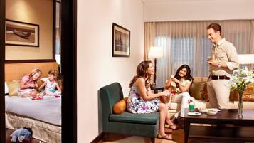 Quarto de hotel com cama, poltrona verde, sofá, mesa de centro e mesa de jantar com um vaso de flores.