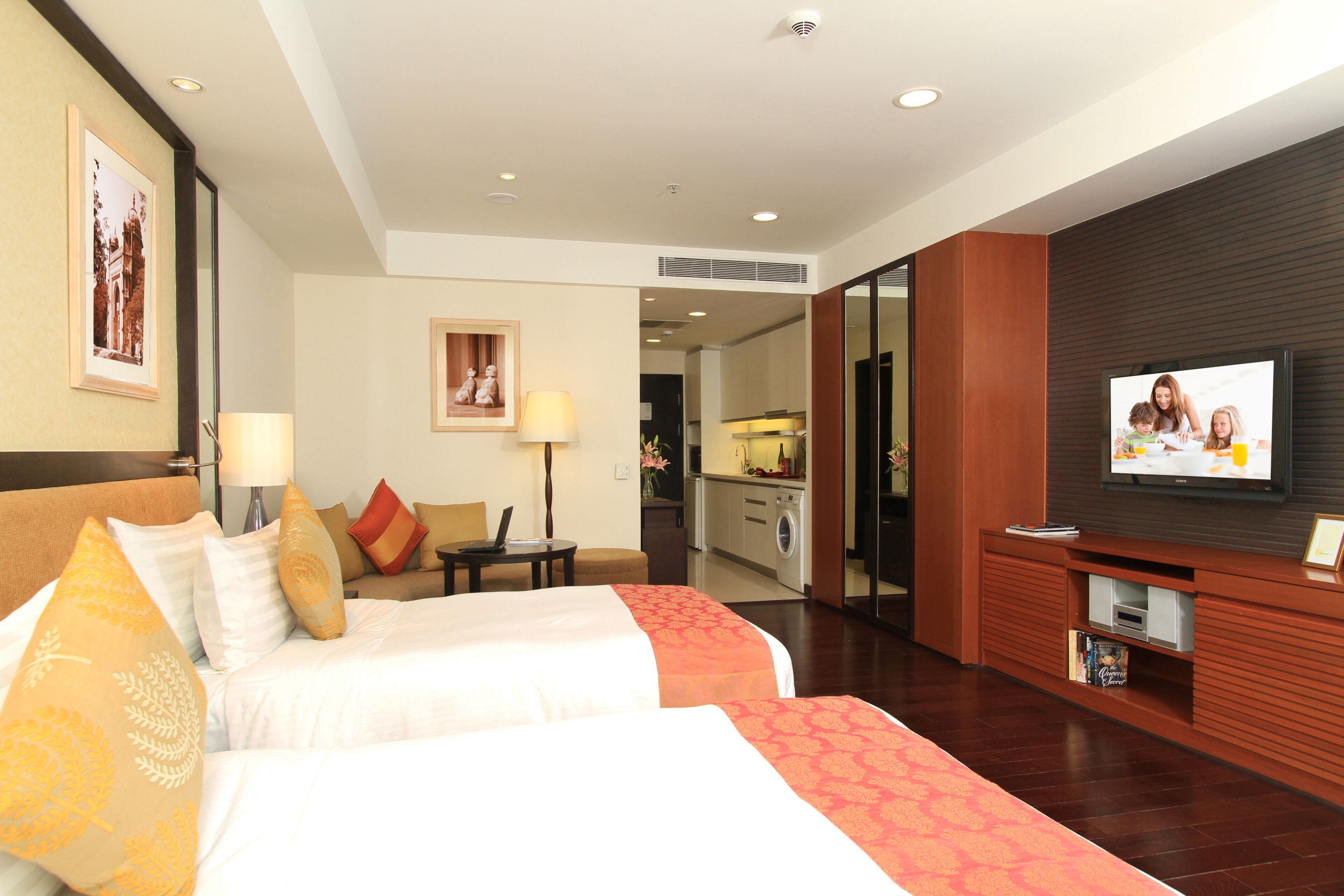 A hotel room with two beds, a TV, a desk, and a kitchenette.