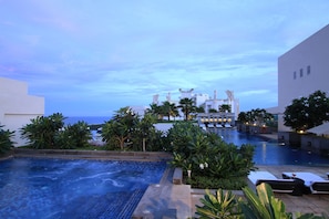 A swimming pool with lounge chairs, surrounded by greenery and a view of a building with a white facade and multiple chimneys.