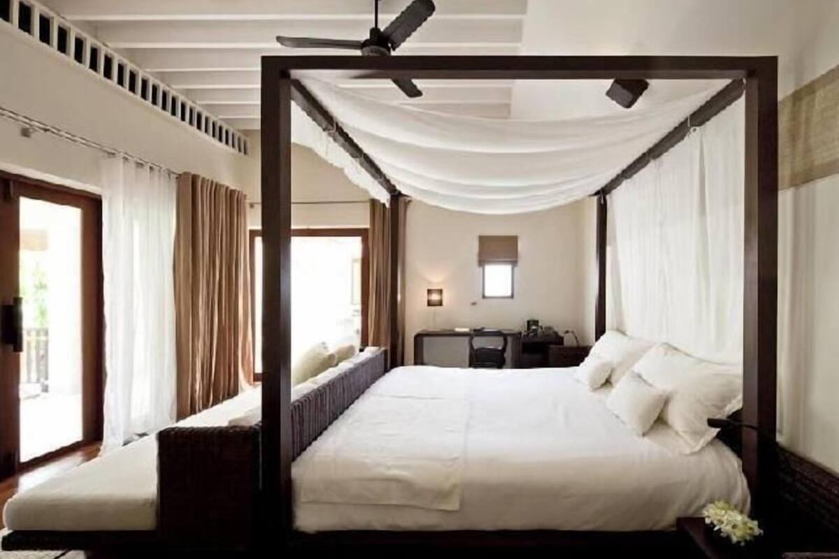 A four-poster bed with a ceiling fan, a desk, and a window with curtains.