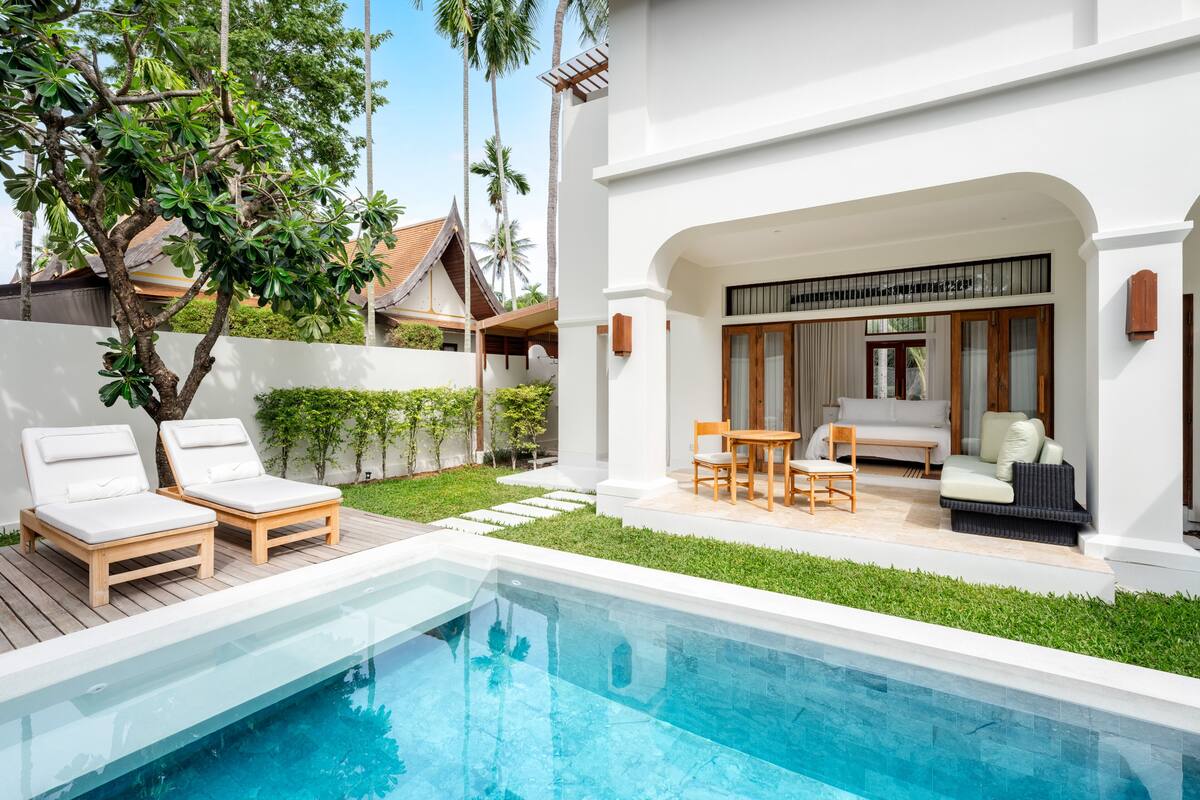 A modern pool area with lounge chairs, a dining set, and a view of a traditional building in the background.