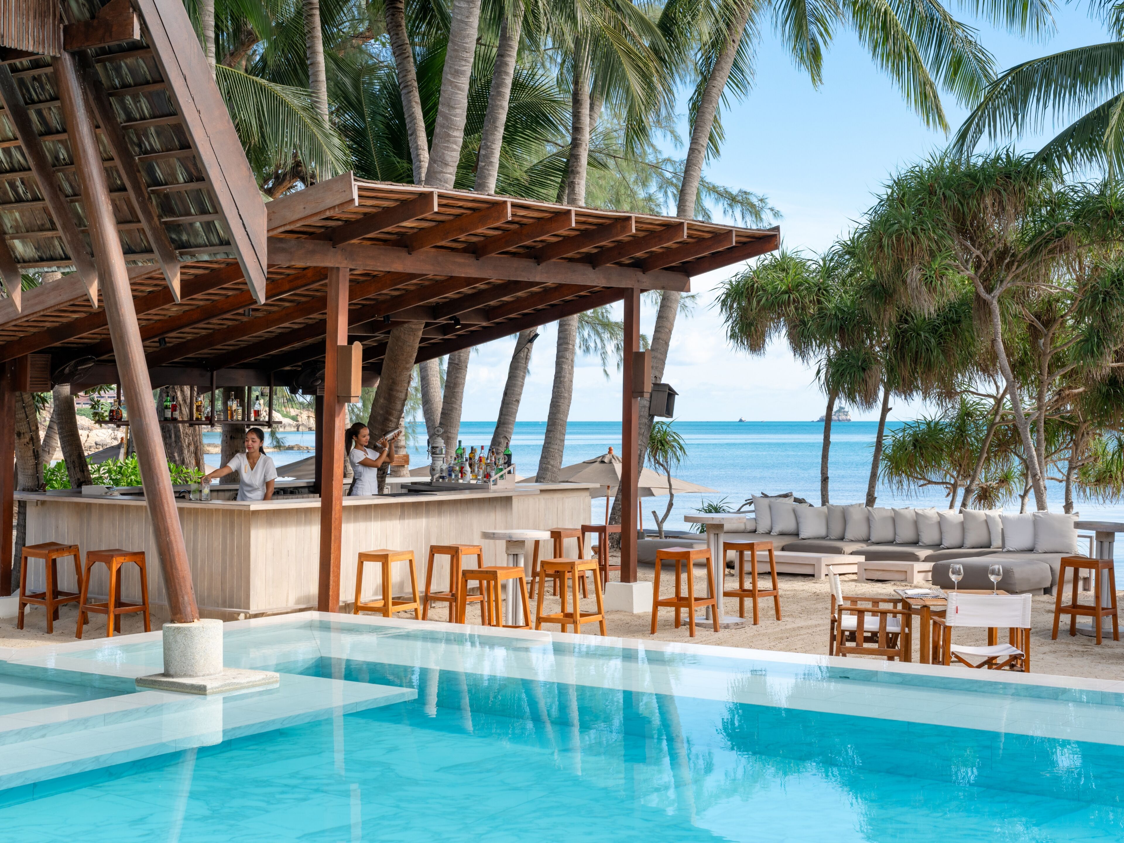 Un bar junto a la piscina con un barman, sillas de madera y una zona de estar con un sofá y mesas.