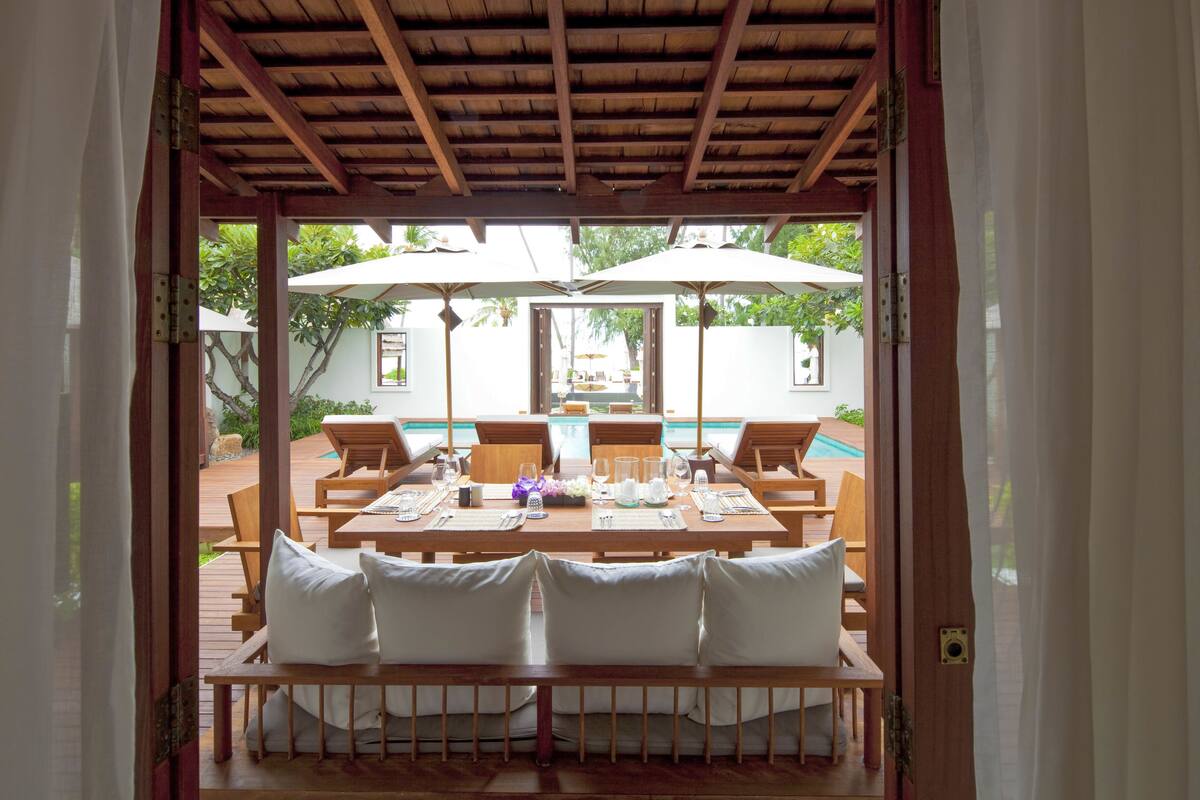 An outdoor dining area with wooden tables and chairs, umbrellas, and a pool in the background.