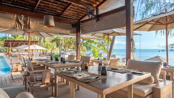 Un área de comedor al aire libre con mesas y sillas de madera, sombrillas y vista al océano.
