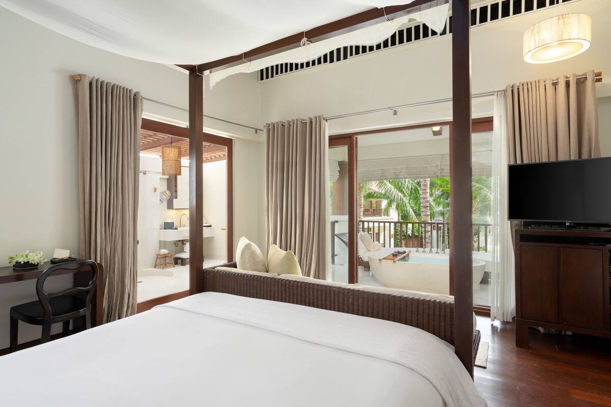 A hotel room with a large bed, a desk with a chair, a TV, and a balcony with a pool view.