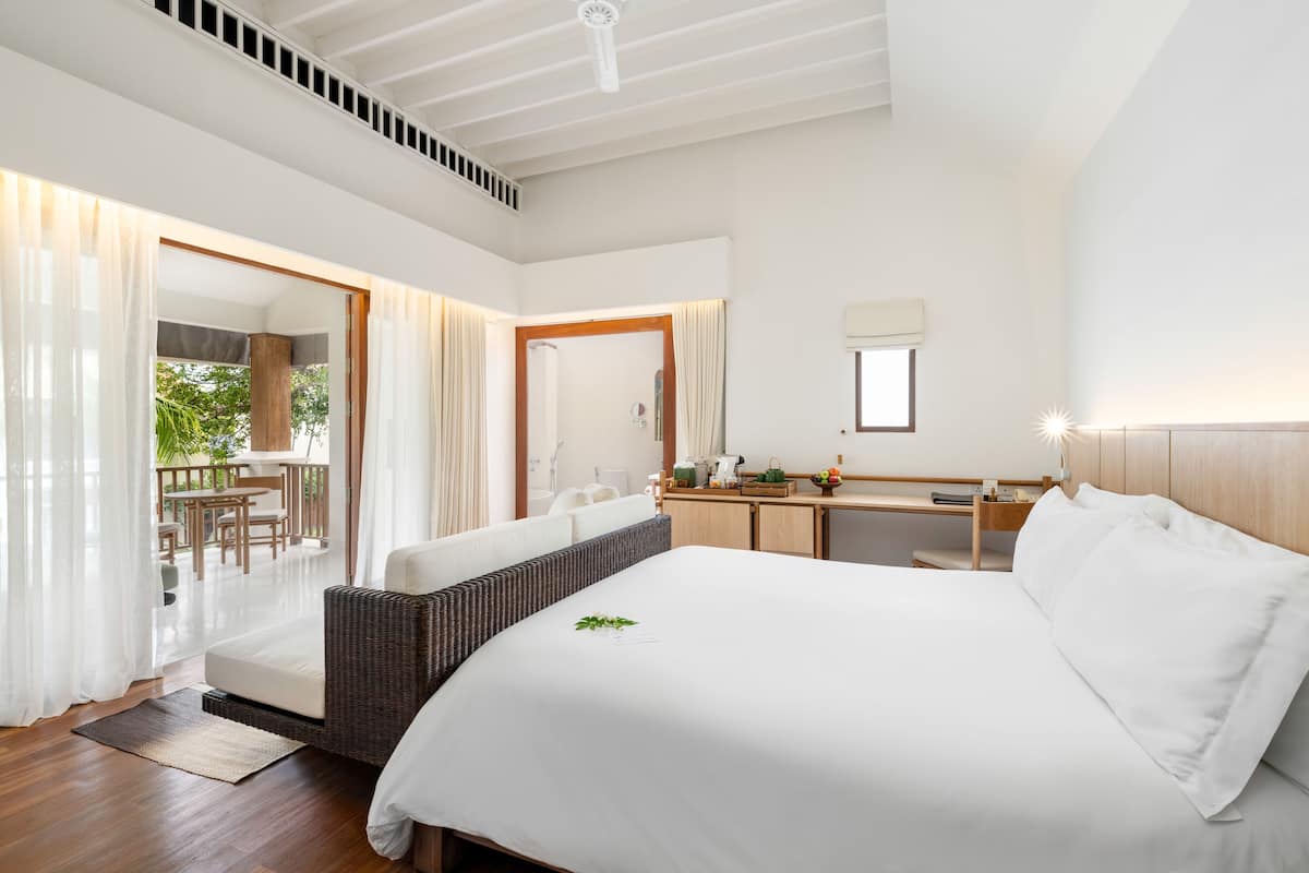 A spacious bedroom with a large bed, a wooden headboard, a bedside table, and a view of a balcony with outdoor furniture.