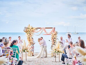 Ceremonia de boda en la playa con una pareja besándose bajo un arco floral.