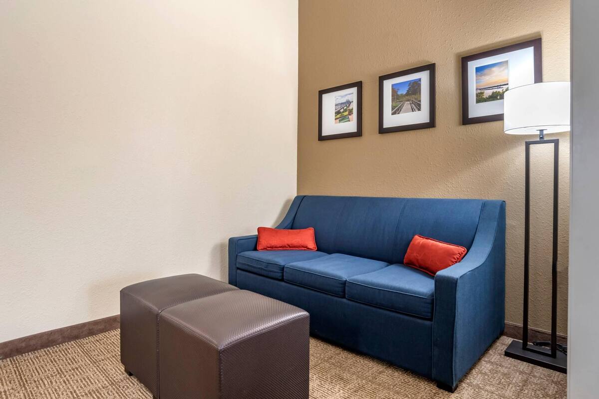 A room with a blue sofa, a brown ottoman, and a floor lamp. There are three framed pictures on the wall.