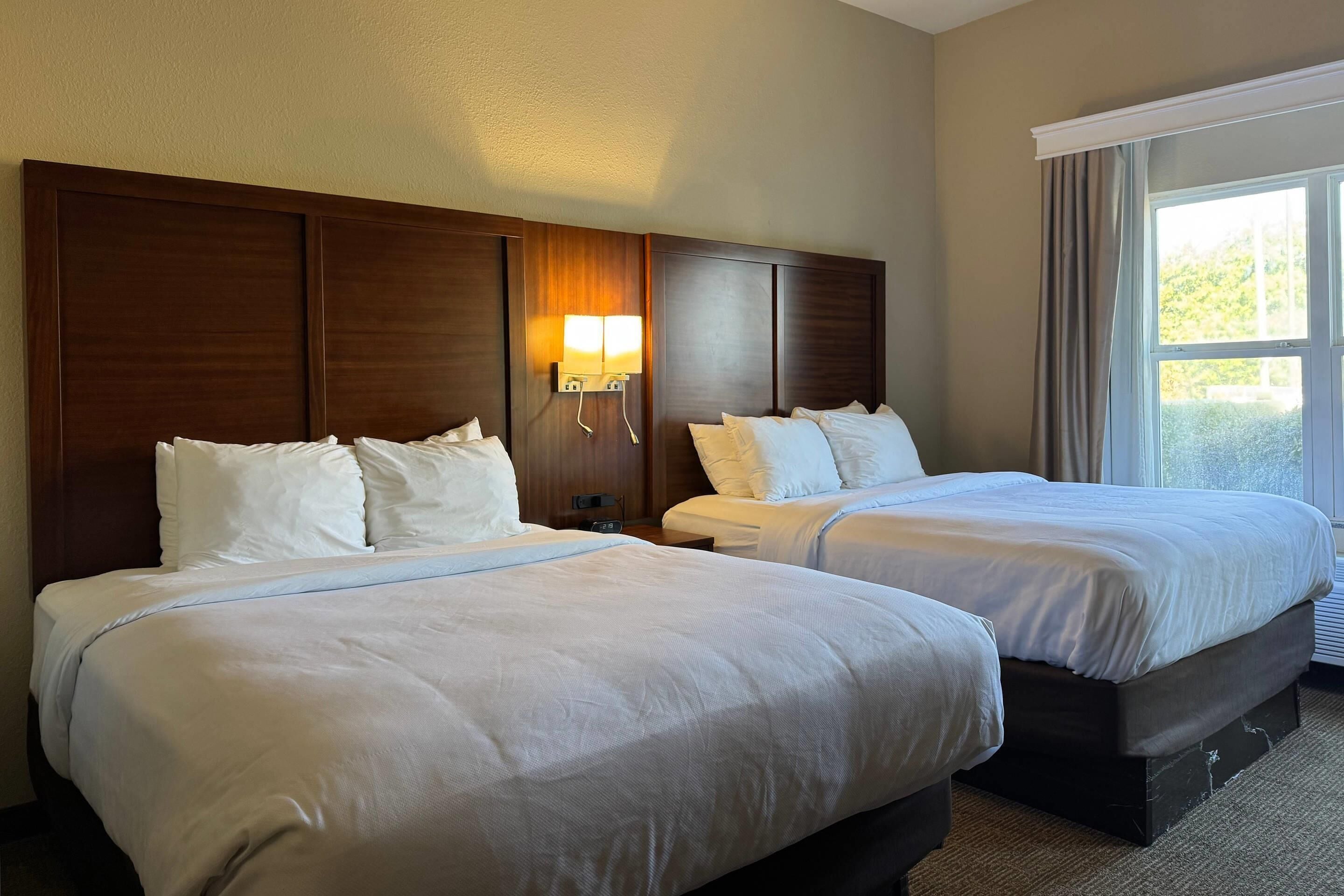 Une chambre d’hôtel avec deux lits, une tête de lit en bois, une table de chevet avec une lampe et une fenêtre avec des rideaux.