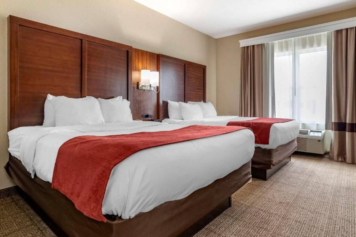A hotel room with two beds, a wooden headboard, a wall-mounted light, and a window with curtains.