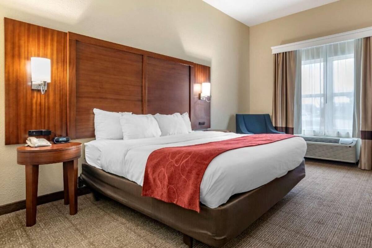 A hotel room with a large bed, a bedside table with a phone, a chair, an air conditioning unit, and a window with curtains.