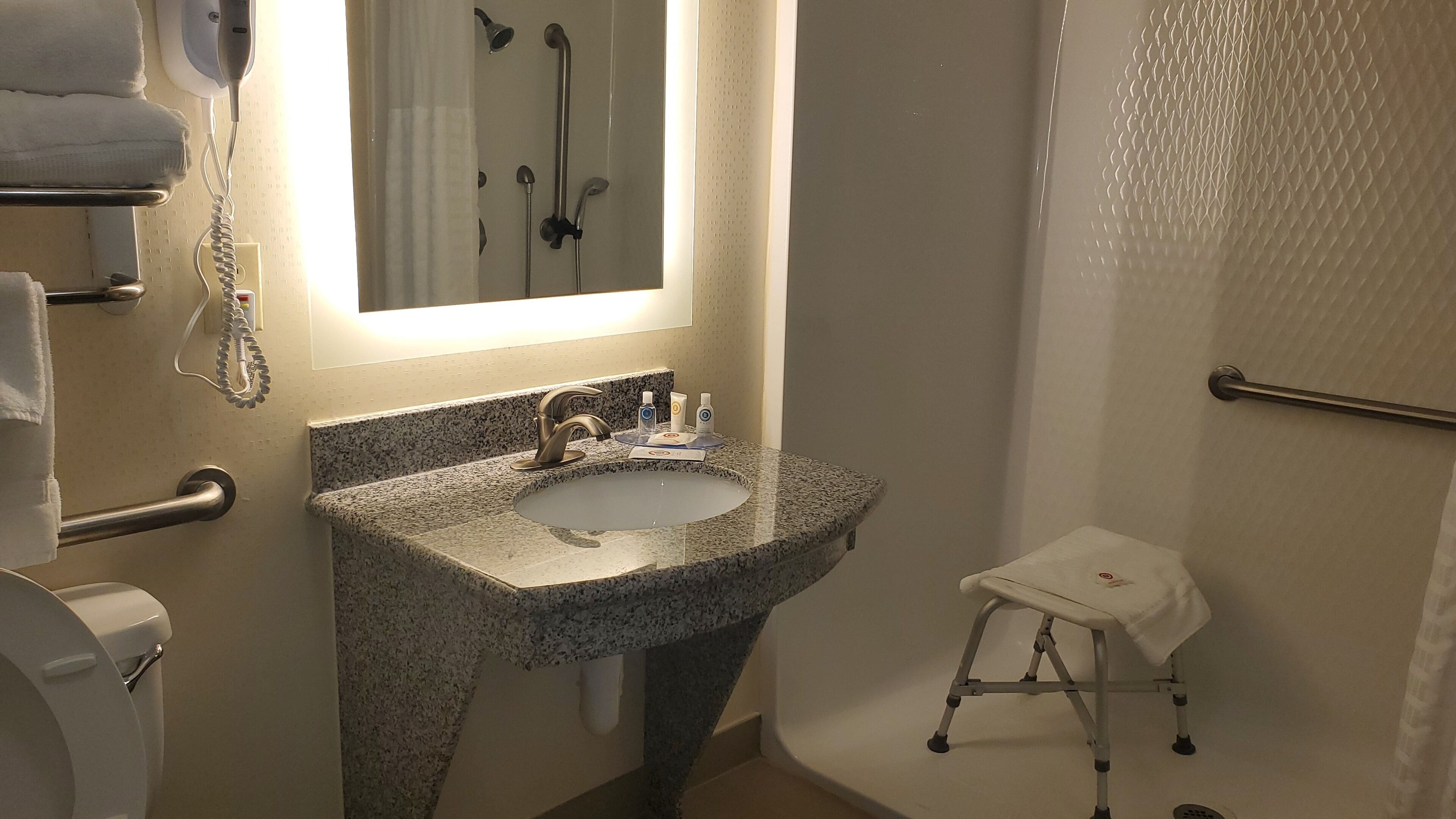 A bathroom with a granite sink, a mirror, a handheld showerhead, a towel rack, and a small stool.