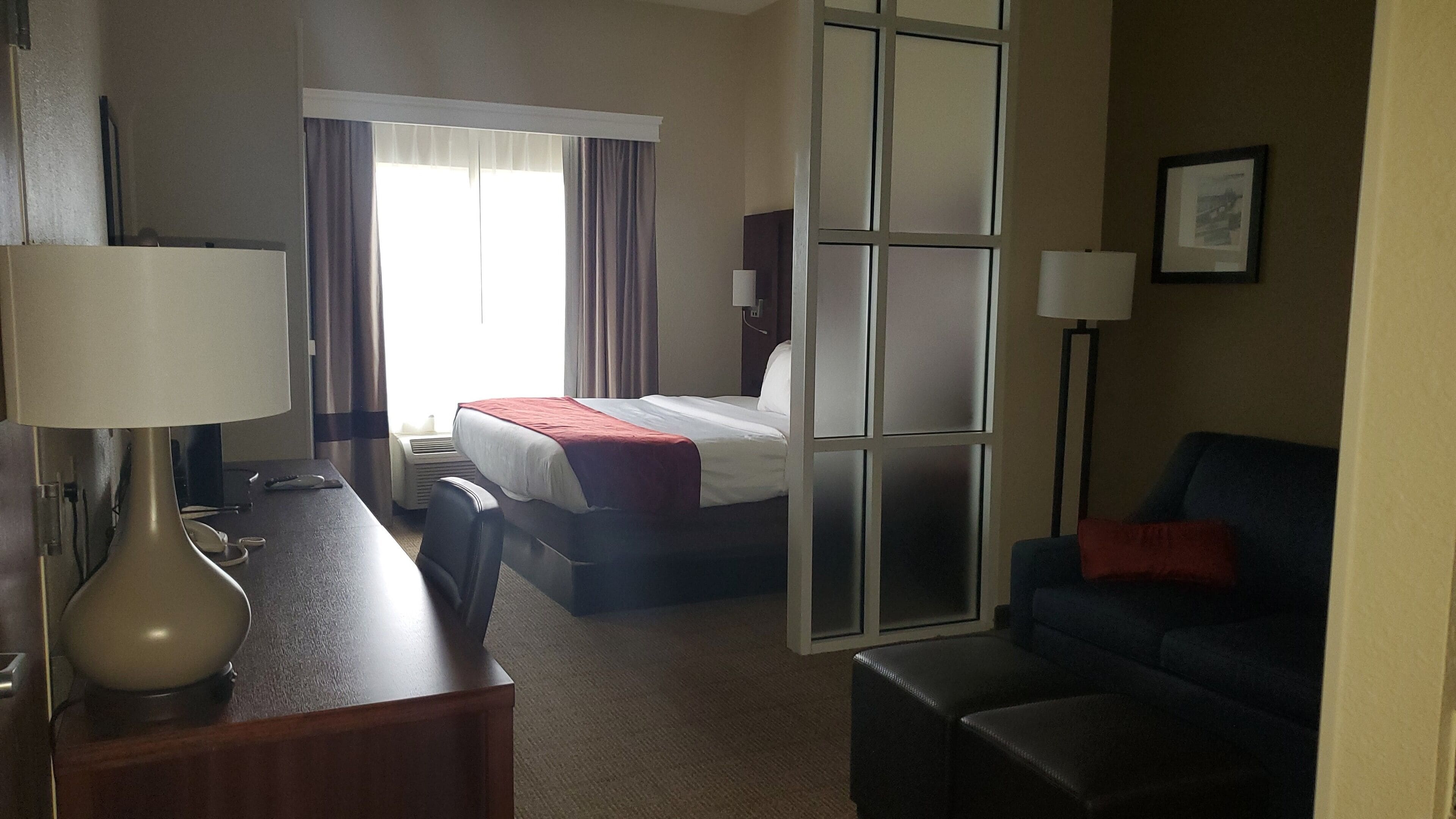 A hotel room with a bed, a desk with a lamp, a chair, a sofa, and a window with curtains.