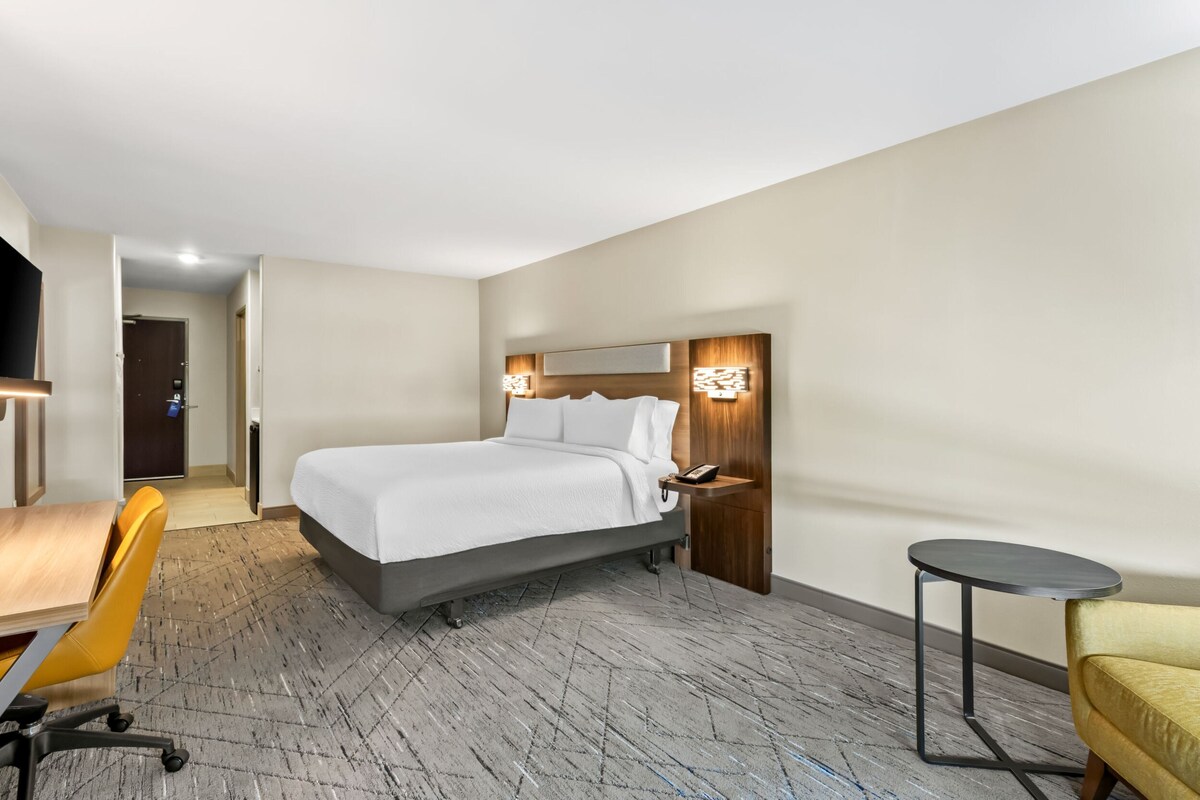 A hotel room with a bed, bedside table, chair, desk, and a small round table.