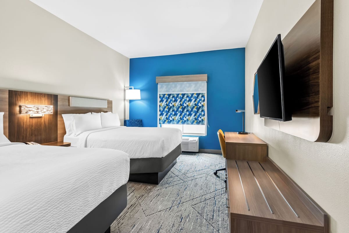 A hotel room with two beds, a desk, a TV, and a window with patterned curtains.